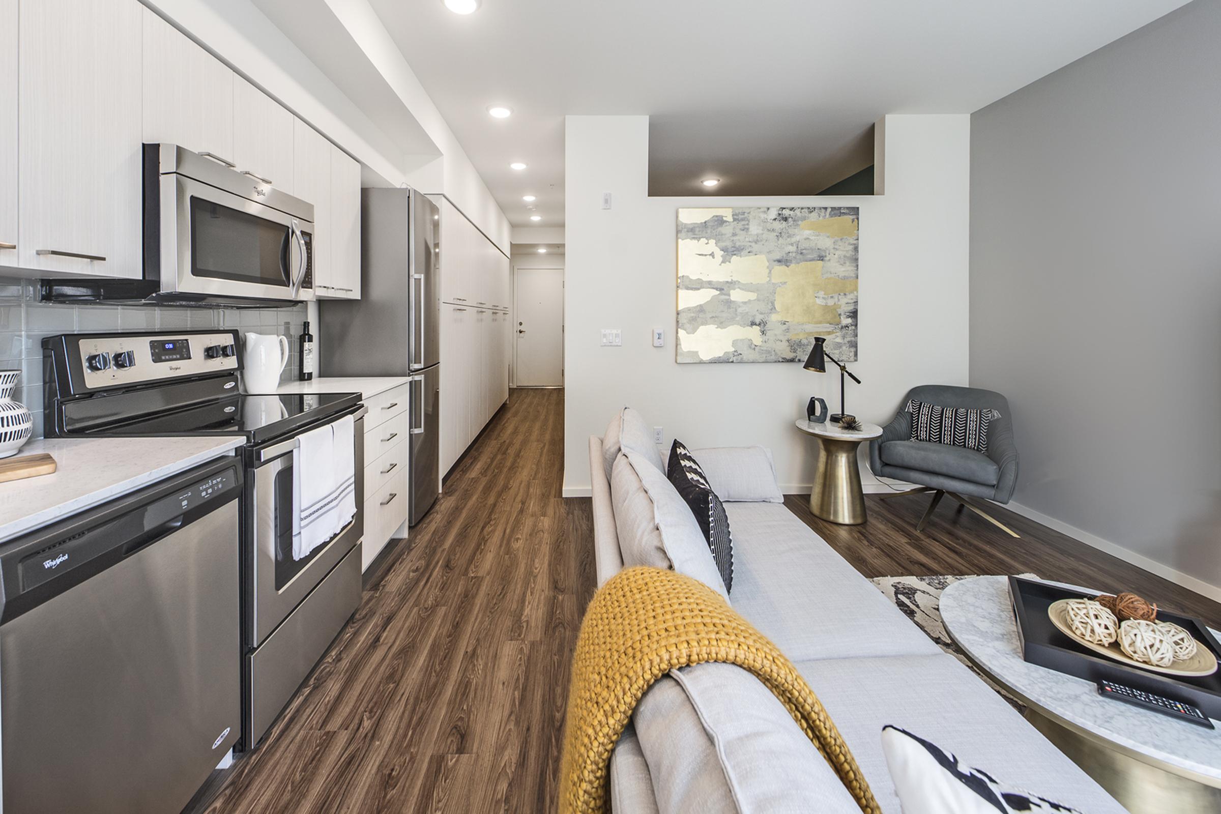 A modern kitchen and living area featuring stainless steel appliances, light wood cabinetry, and a cozy sofa with yellow throw.