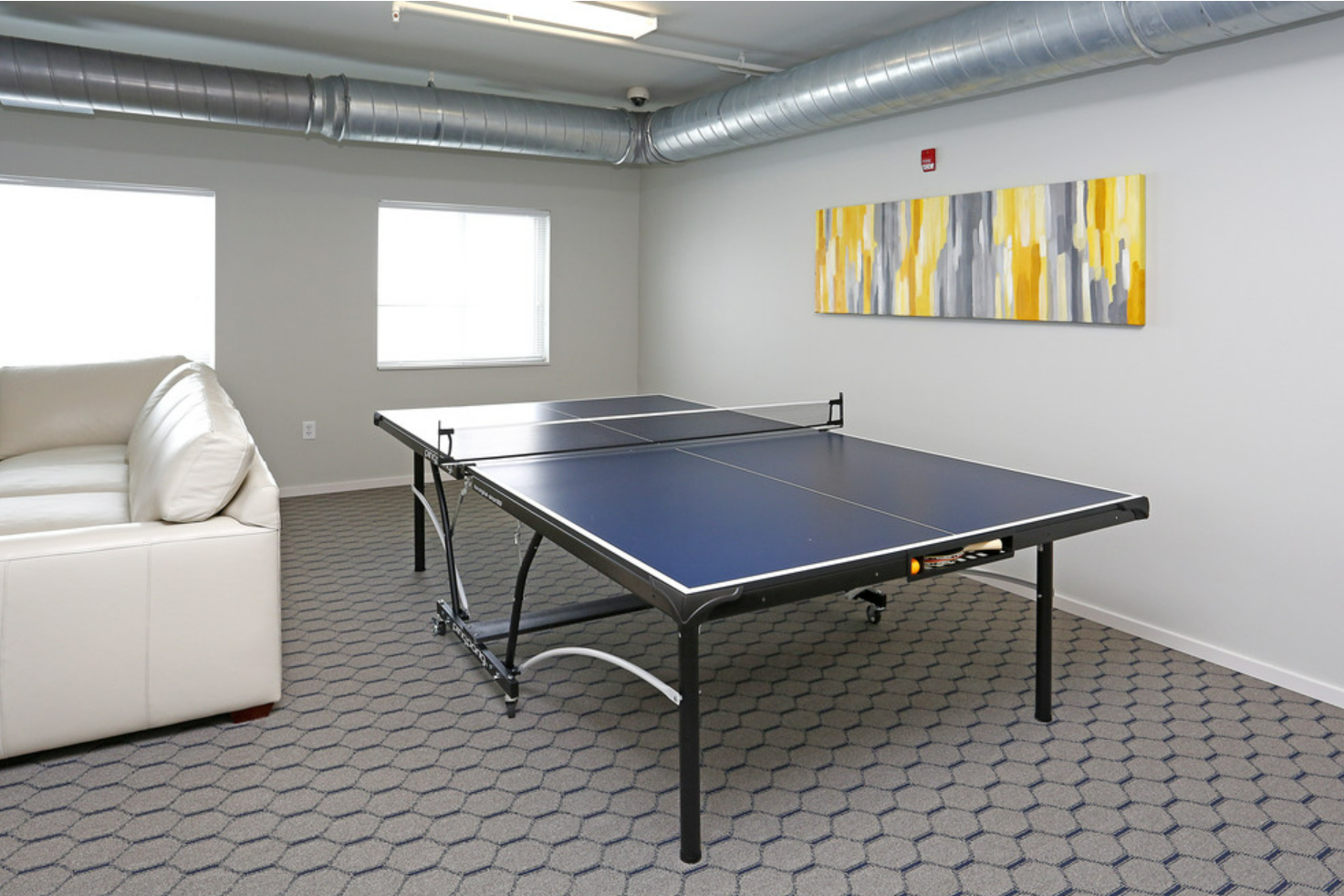 A bright, modern room featuring a table tennis table in the center, with a white sofa on one side. The walls are light gray, and there are two windows allowing natural light. A colorful abstract painting hangs above the table, adding a touch of decor to the space. The floor is covered with hexagonal-patterned carpet.