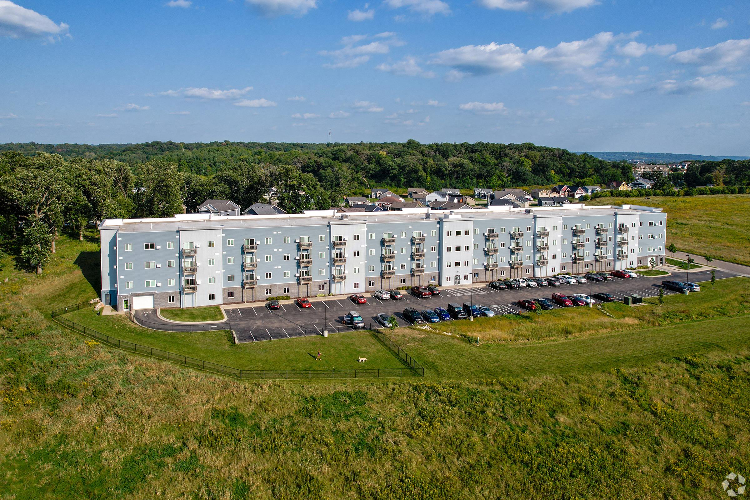 A modern multi-story residential building with light blue siding, situated on a green, grassy area. The building features balconies and a parking lot in front, with several cars parked. In the background, there are trees and distant residential houses under a partly cloudy sky.
