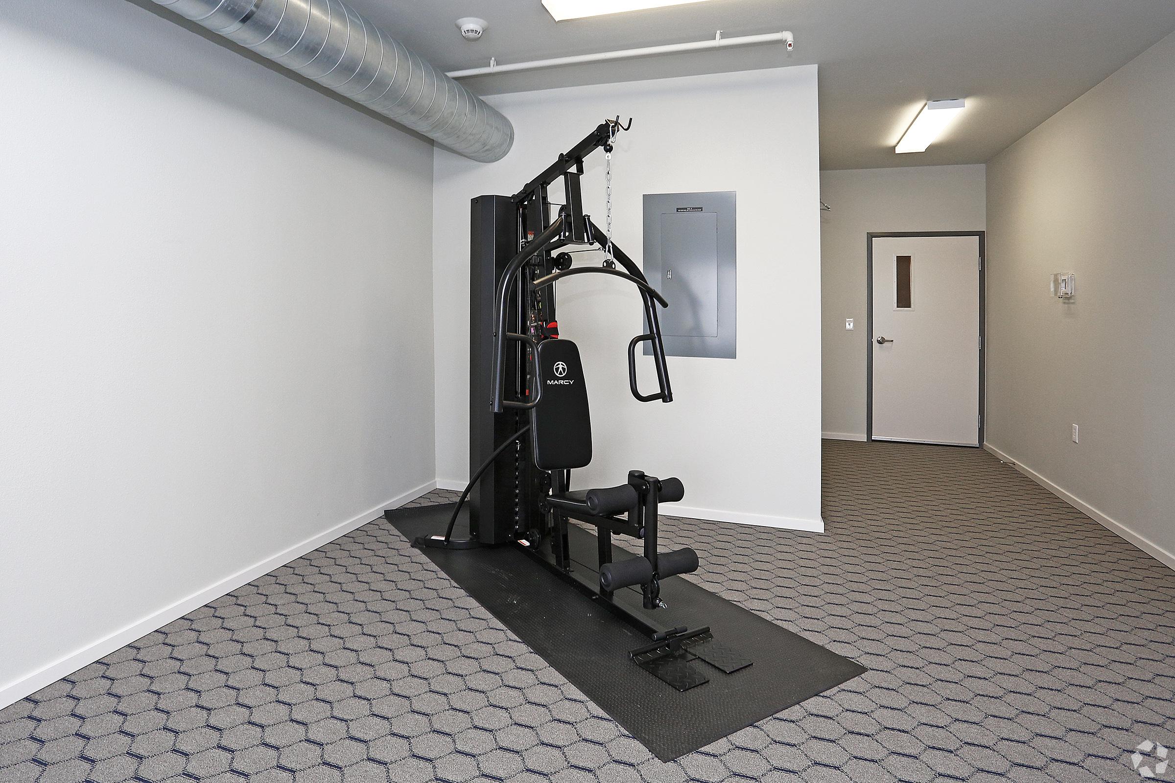 A small, simple gym space featuring a multi-gym machine on a black mat. The room has light gray walls, a door with a small window, and an electrical panel on the wall. A ventilation duct runs along the ceiling, and the floor is covered with a patterned gray carpet.