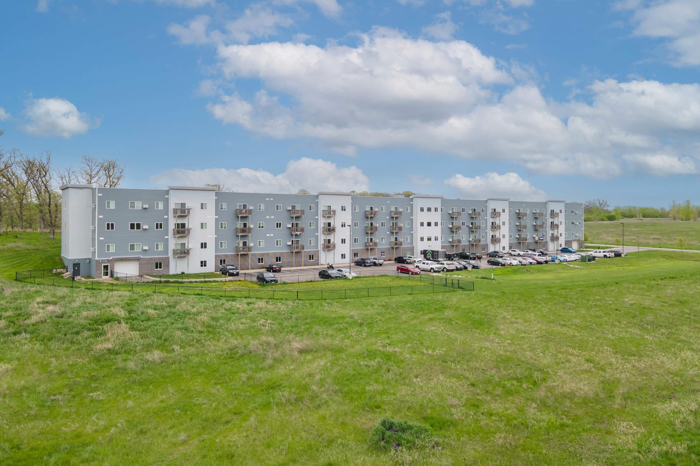 A modern multi-story apartment building in a grassy area, featuring grey and white exteriors. Vehicles are parked in front, with trees and a blue sky in the background, scattered with fluffy clouds. The scene conveys a suburban residential environment.
