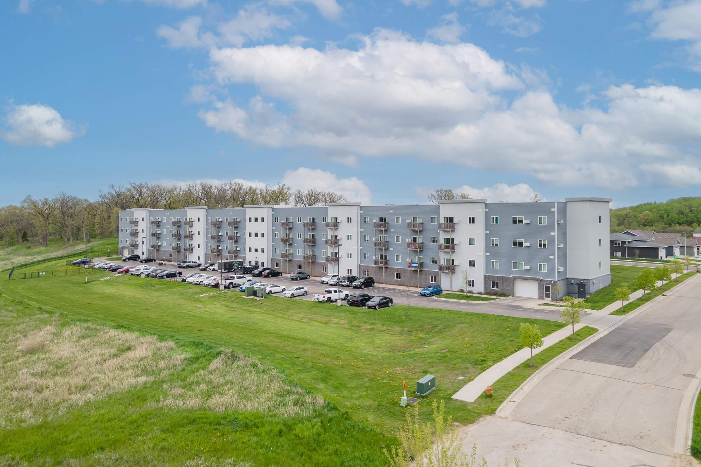 A modern multi-story apartment building with a gray exterior, surrounded by green grass and trees. The building features multiple balconies and a parking lot filled with vehicles. The area appears residential, with a blue sky and fluffy clouds in the background.