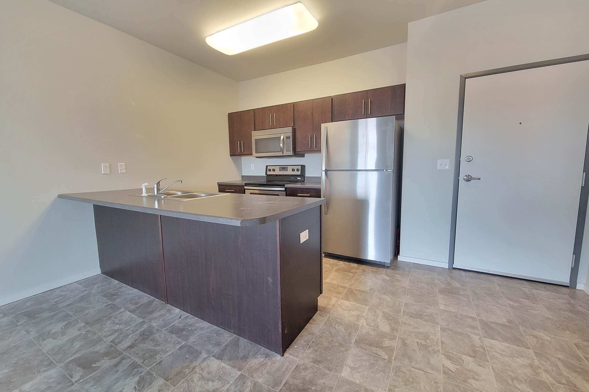 Modern kitchen featuring dark wood cabinetry, stainless steel appliances including a fridge and microwave, and a central island with a sink. The flooring is tile, and there is a large overhead light fixture. A door is visible on the right side, leading to another area.
