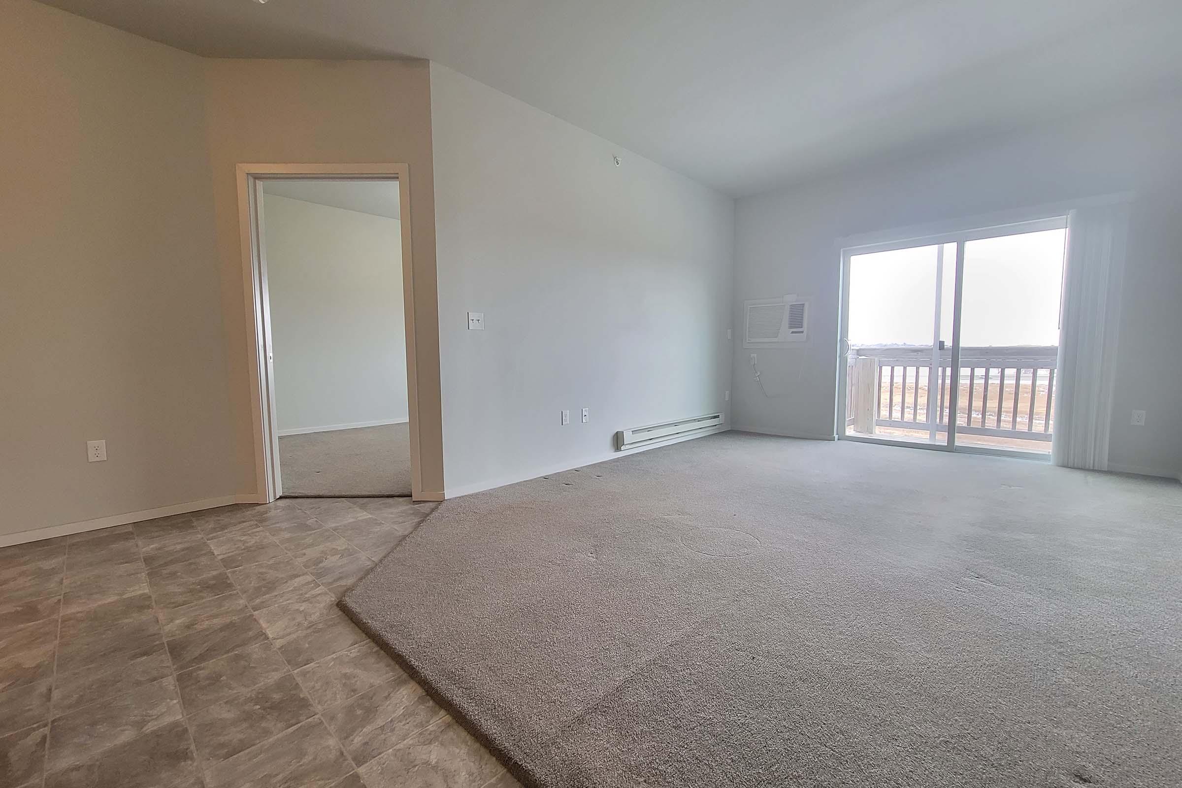 Empty room featuring light gray walls and a large window with sliding doors leading to a balcony. There is a door on the left side leading to another space, and an air conditioning unit mounted on the wall. The floor is covered with light-colored carpet, and the room is well-lit.