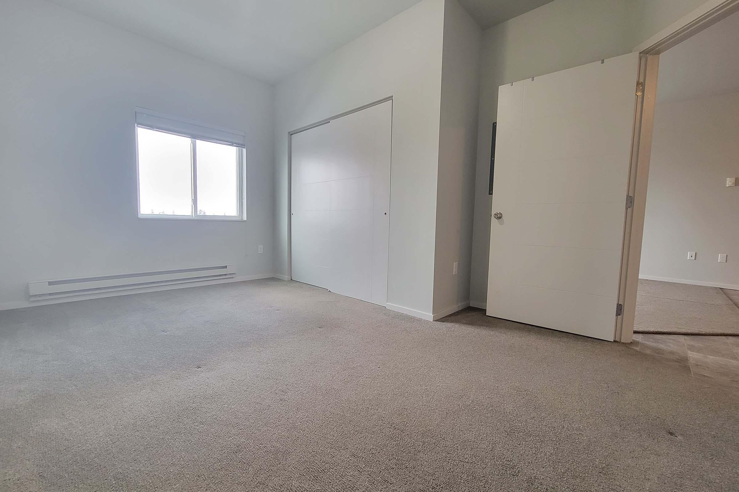 A bright, empty room with light-colored walls and carpeting. There is a window on the left side allowing natural light to enter, a closet door on the back wall, and an open door leading to another room on the right. The space appears clean and unoccupied, suggesting it is available for new occupants.