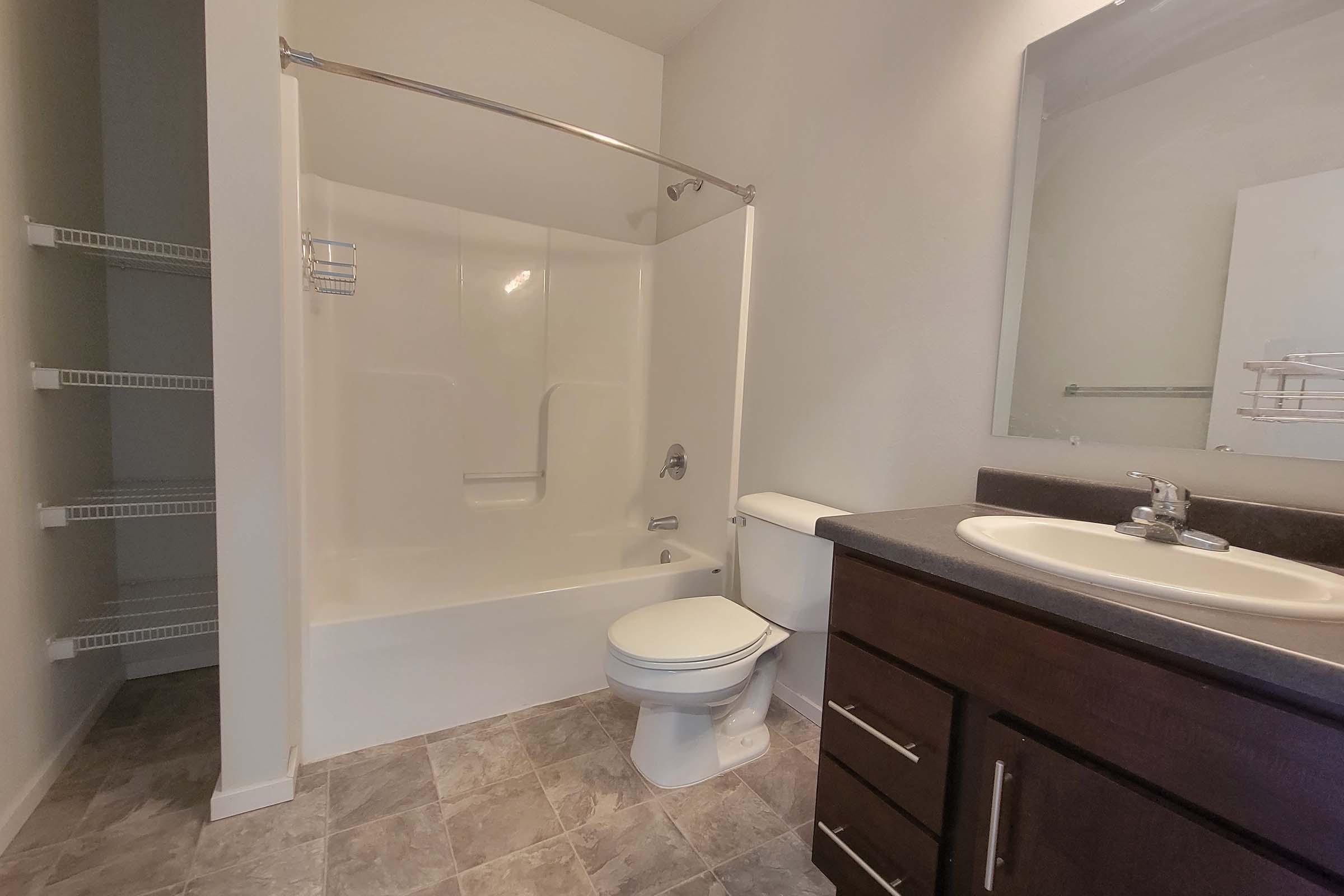 A clean bathroom featuring a white tub and shower combo, a toilet, and a sink with dark wood cabinets. The wall is painted light, and there is a large mirror above the sink. On the left, there are open shelves for storage. The floor has a light tile pattern.