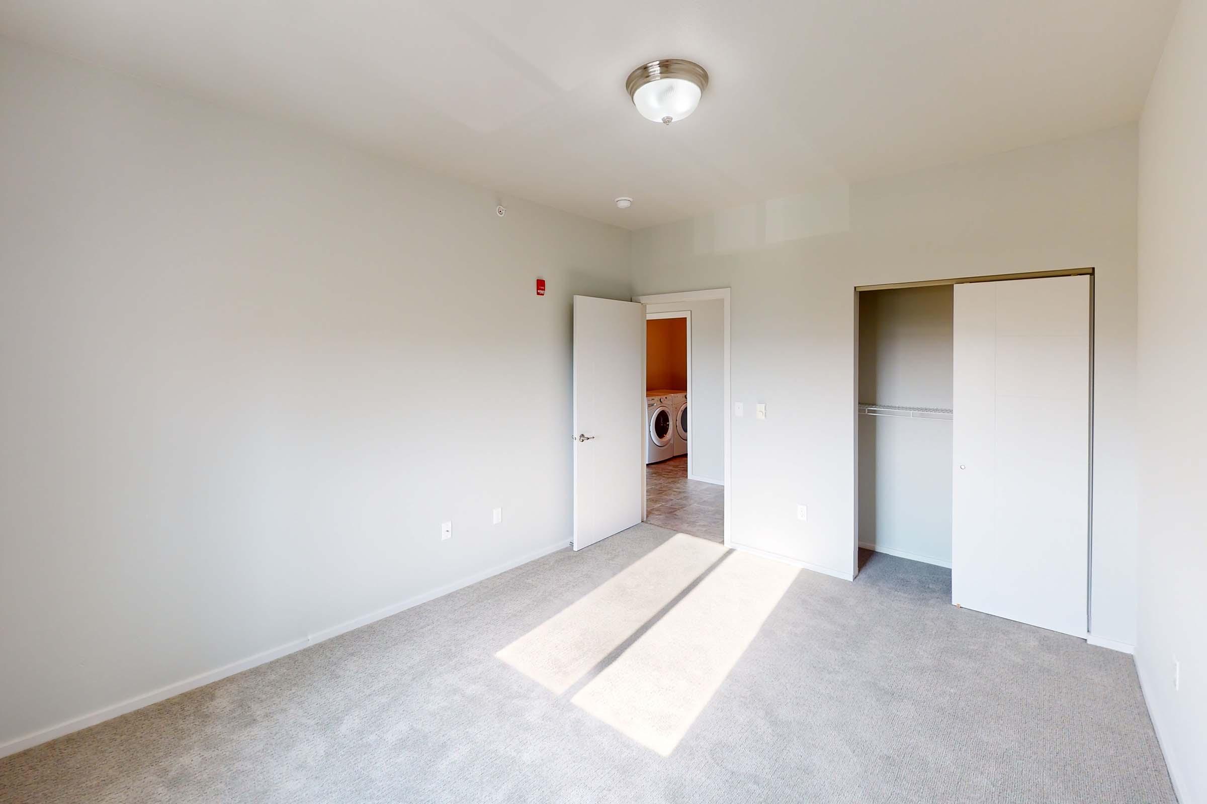 A brightly lit room with light gray walls and a carpeted floor. It features an open door leading to a laundry area, a closet with sliding doors, and a ceiling light fixture. The sunlight casts shadows on the floor, enhancing the spacious feel of the room.
