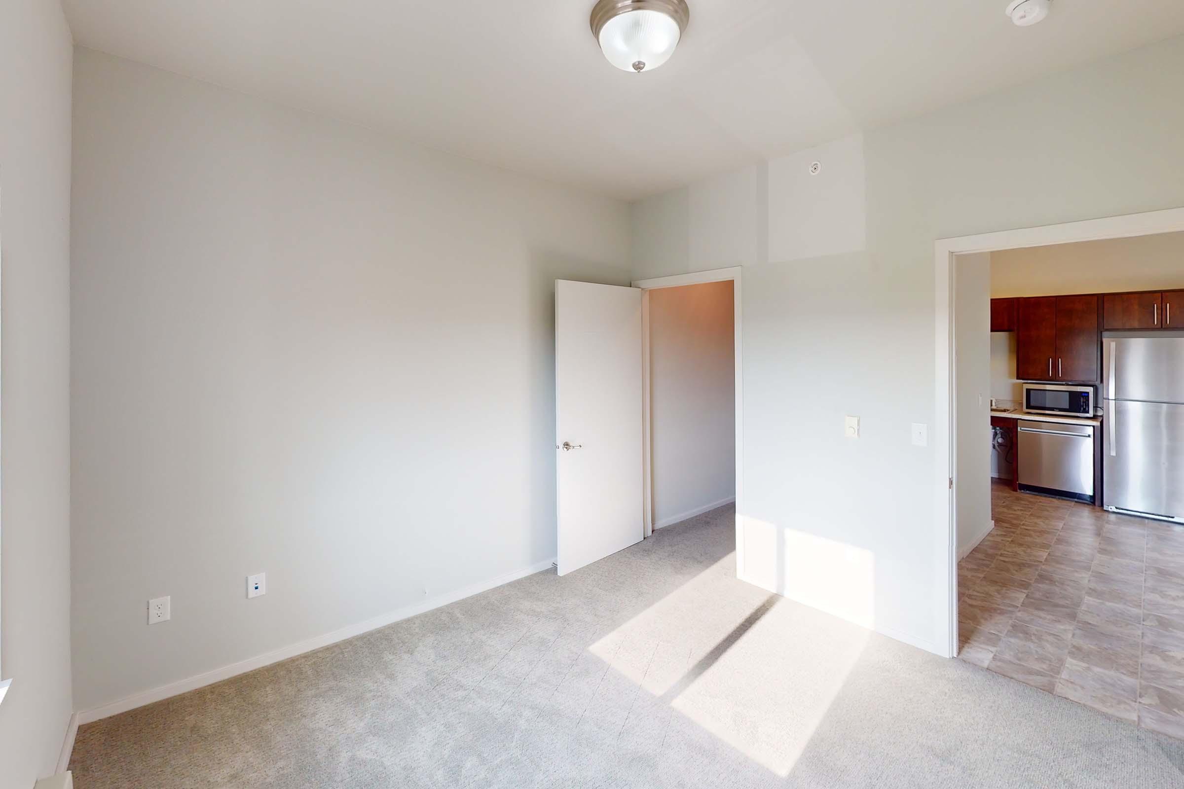 A well-lit room featuring beige carpet and light gray walls. A closed white door leads to another area, likely a kitchen, visible through an open doorway. Sunlight casts shadows on the floor, enhancing the spacious feel.