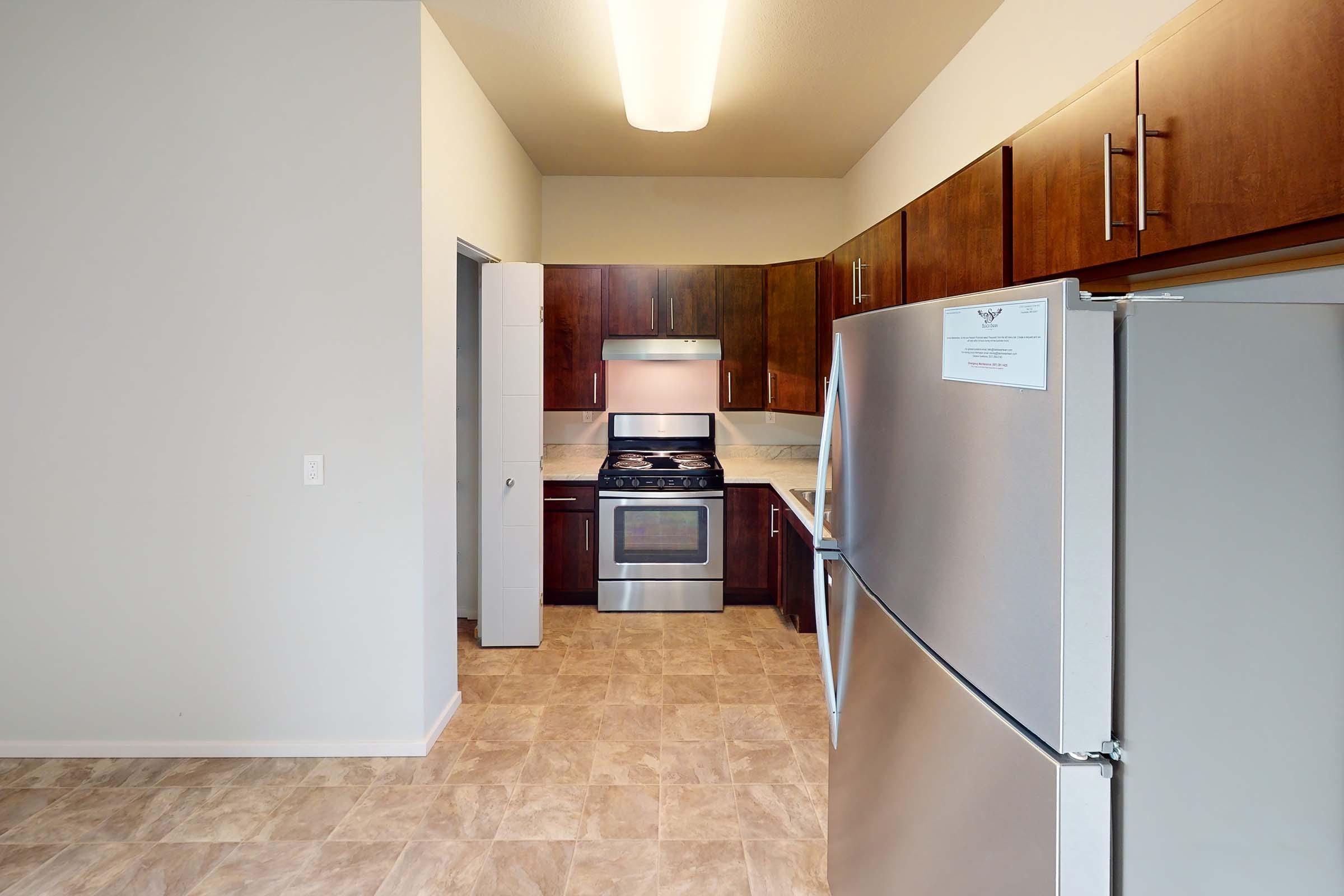 Modern kitchen with wooden cabinets, stainless steel appliances, and a gas stove. The space features tiled flooring and ample lighting, making it bright and inviting. A doorway leads to another room, and the refrigerator is prominently displayed on the right.