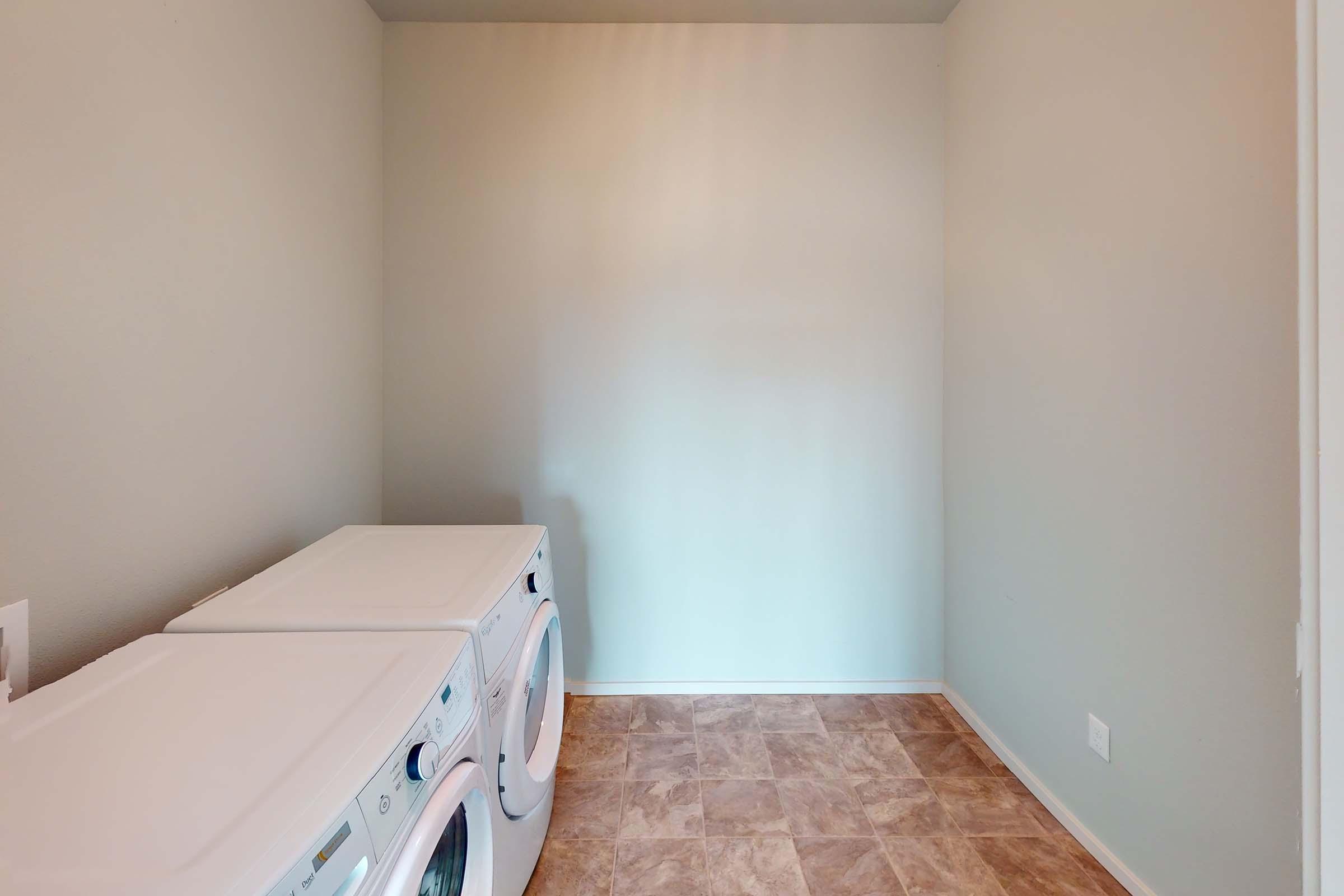 A spacious laundry room featuring white washer and dryer appliances. The walls are painted light gray, and the floor consists of tan vinyl tiles. Natural light fills the room, creating a bright and clean atmosphere.