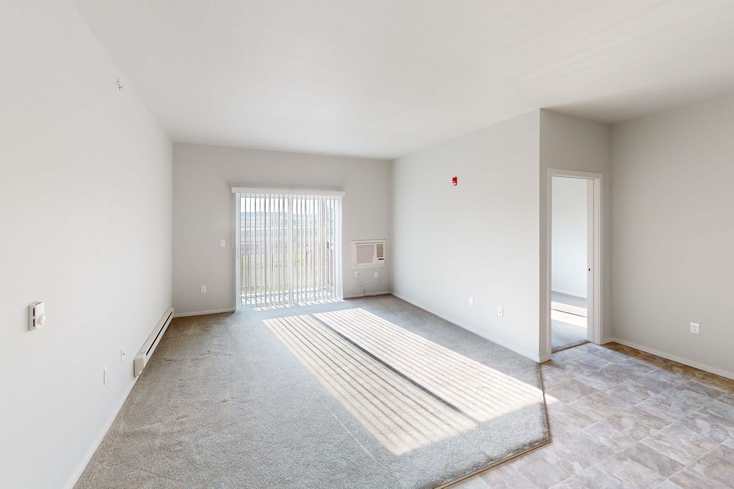 Bright, empty living room with light gray walls and carpet. A large window with vertical blinds lets in natural light, creating shadows on the floor. There is an air conditioning unit on the wall and a doorway leading to another room. The space feels open and is ready for furniture.