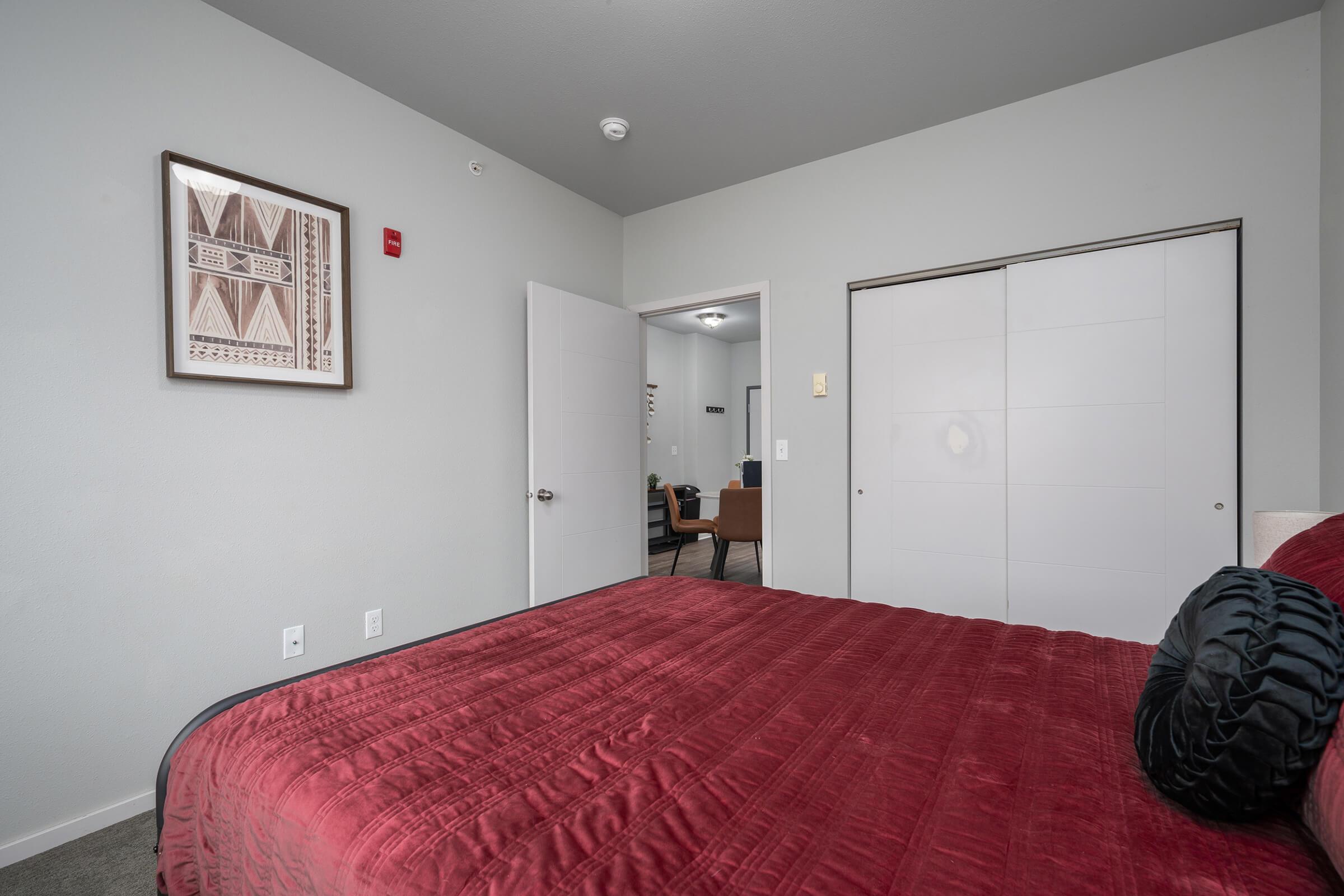 A well-decorated bedroom featuring a large red bedspread, a framed artwork on the wall, and a closet with sliding doors. The room has light gray walls and a doorway leading to another area, which is partially visible in the background. A cozy armchair can be seen through the open door.