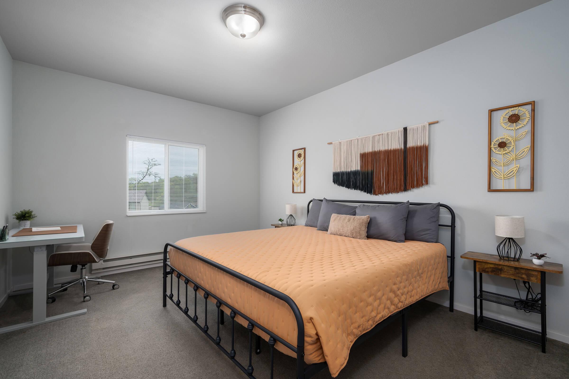 A cozy bedroom featuring a queen-sized bed with an orange quilt, a nightstand with a lamp, a small wooden table, and a desk with a chair. The room has white walls, a window with light filtering through, and decorative wall hangings. Perfect for a calm and inviting atmosphere.