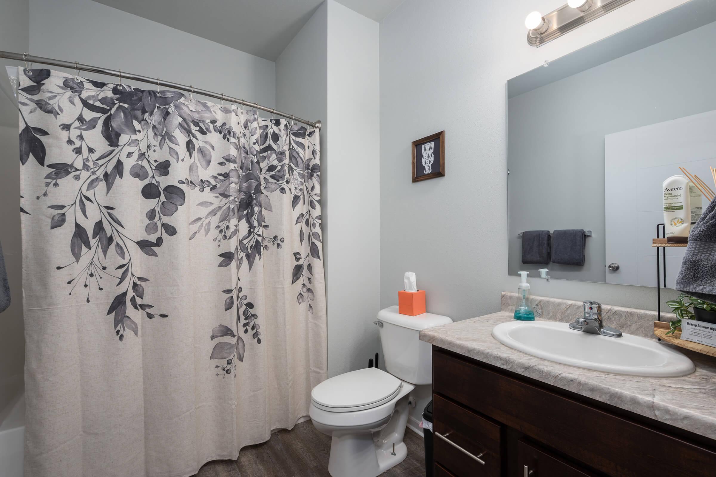 A modern bathroom featuring a light-colored shower curtain with a floral design, a white sink with a dark wooden cabinet, a toilet, and a mirror. The walls are painted in a neutral tone, and there are decorative items like a small plant and toiletries on the countertop.