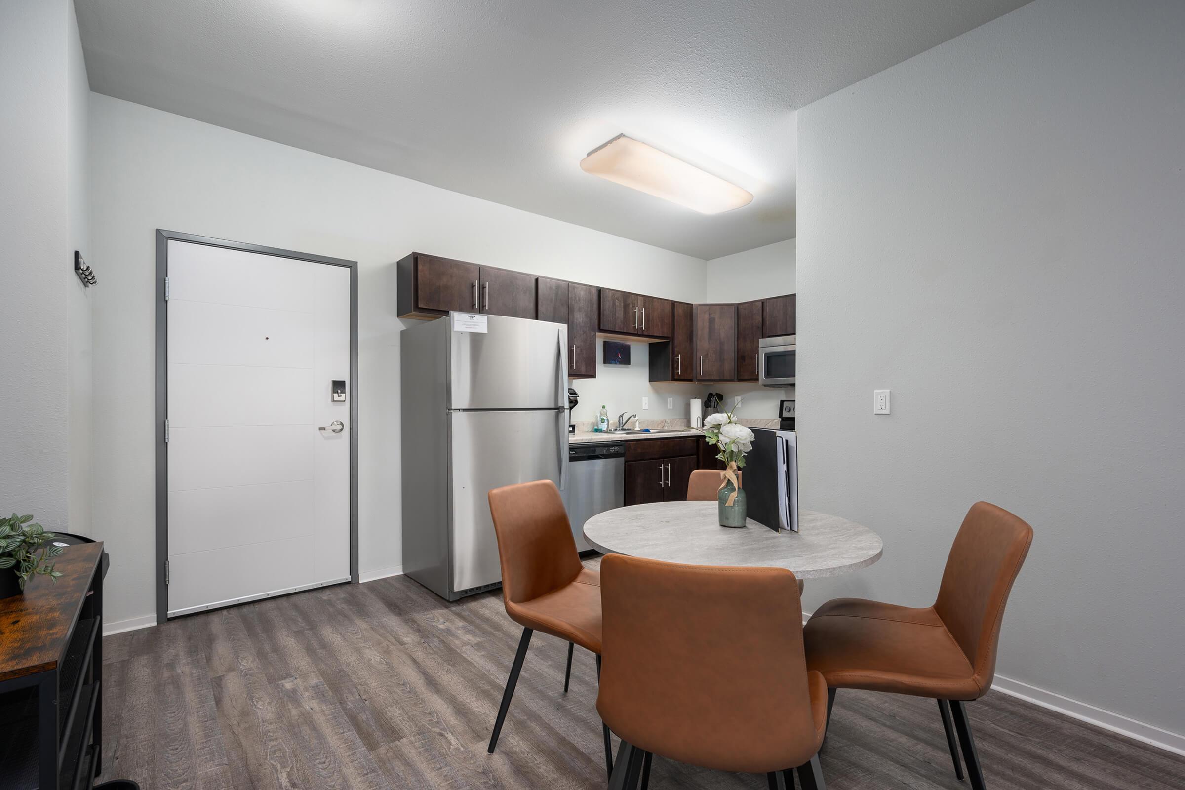 Modern kitchen and dining area featuring a round table with four brown chairs, a refrigerator, a microwave, and dark wood cabinets. A door leads to another room, and a vase with flowers is on the table. The flooring is a light wood laminate, creating a bright and inviting atmosphere.