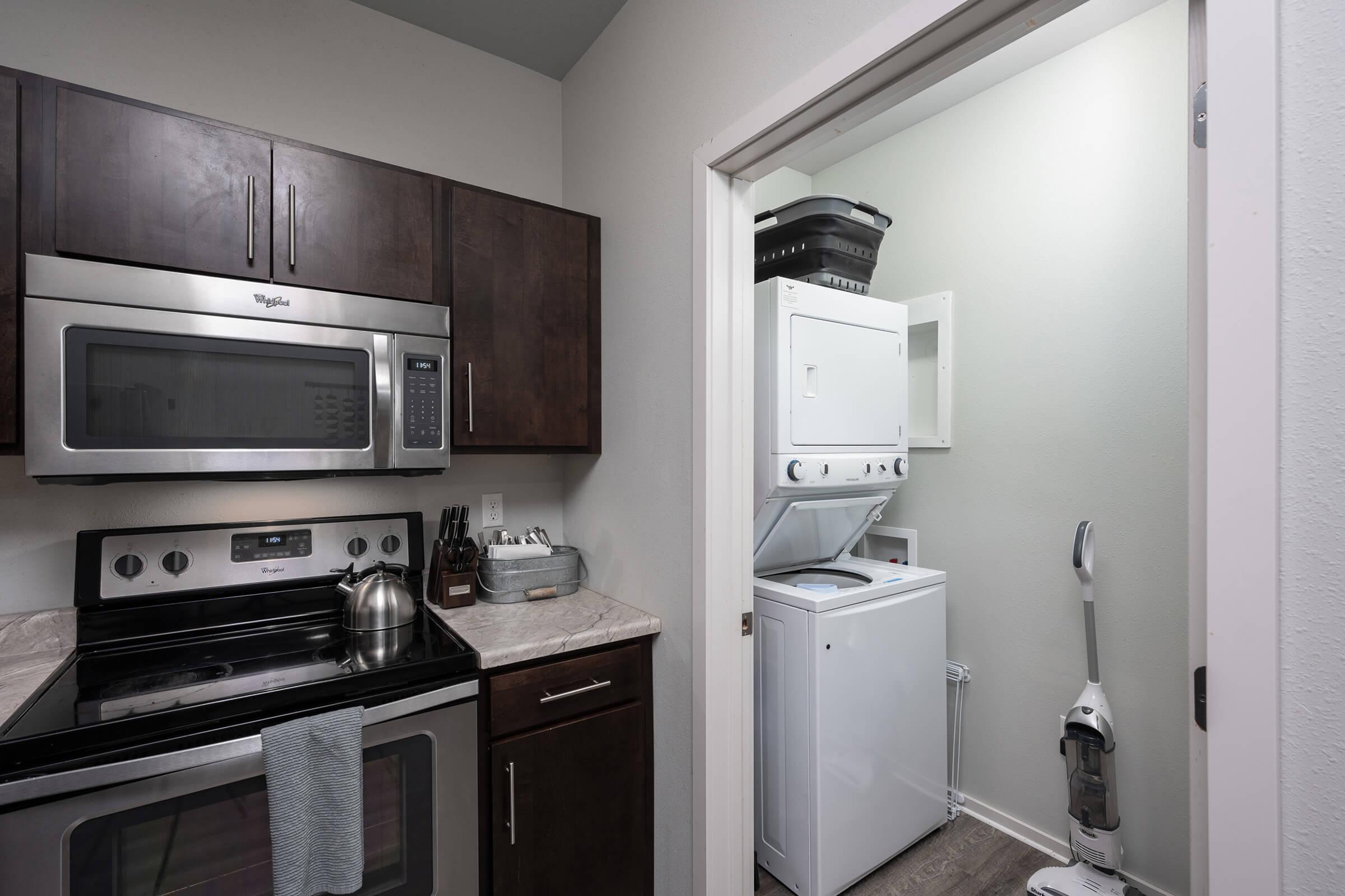 A modern kitchen featuring dark wood cabinets, stainless steel appliances including a microwave and oven, and a laundry area visible through an open door with a washing machine, dryer, and cleaning supplies. The countertops are light-colored, complementing the overall design.