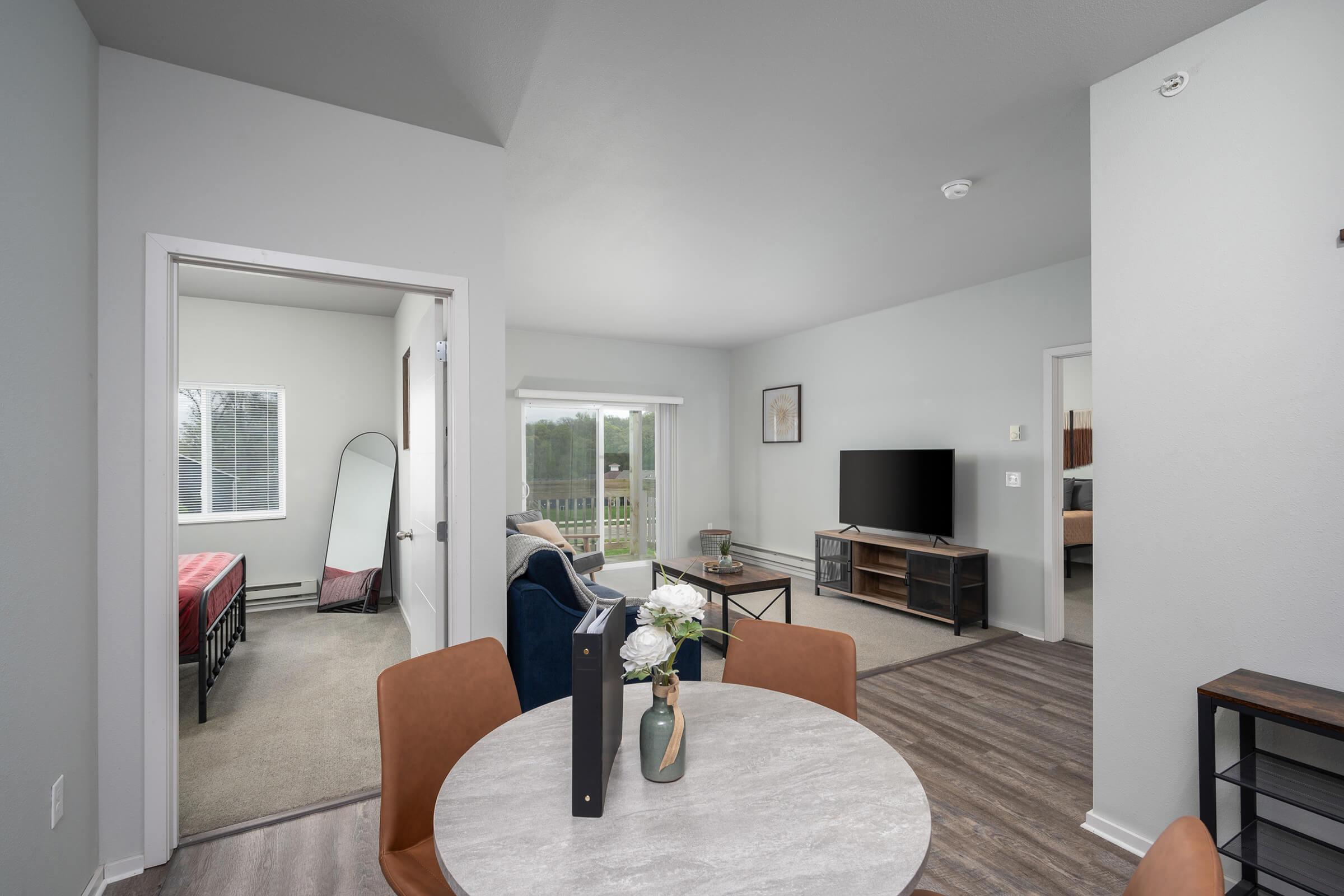 A modern living space featuring a dining table with chairs, a cozy blue couch, a television unit, and an open doorway leading to a bedroom. Natural light fills the room through large windows, and decorative elements like a vase with flowers add a welcoming touch.