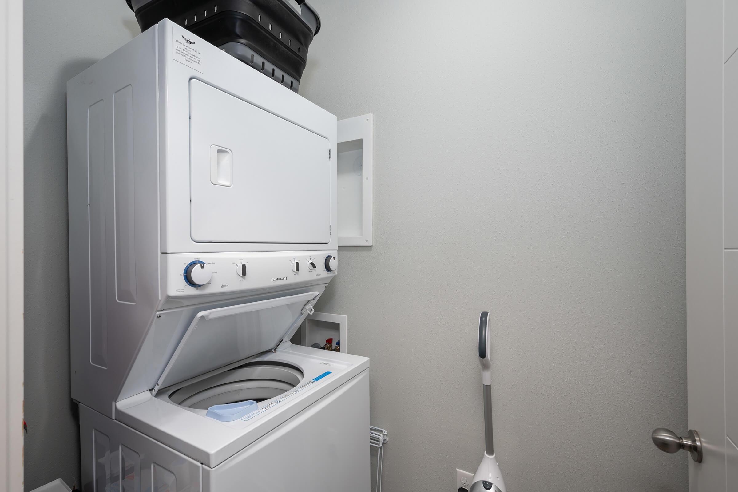A compact laundry area featuring a stacked washer and dryer unit in white, with a black storage basket on top. A steam mop is positioned nearby against the wall, and the walls are painted a light gray color. The entrance to the space is visible, with a modern door handle.