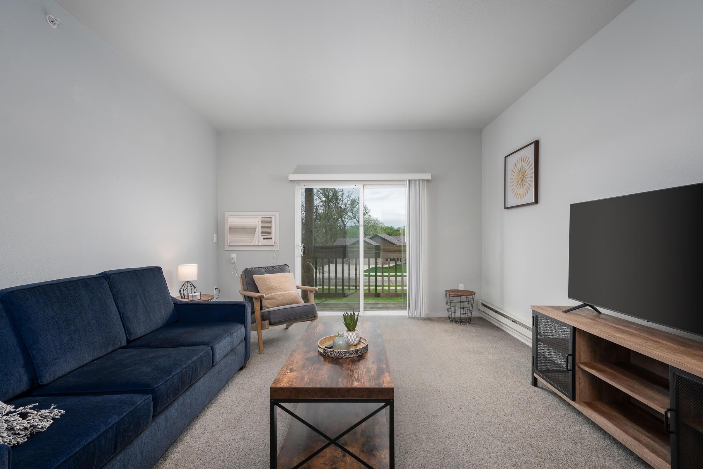 Modern living room featuring a navy blue sofa and a wooden coffee table. An armchair is beside the sofa, and a television sits on a stand. A window with sliding glass doors leads to an outdoor view, while an air conditioning unit is mounted on the wall. The decor includes a wall art piece and a potted plant.