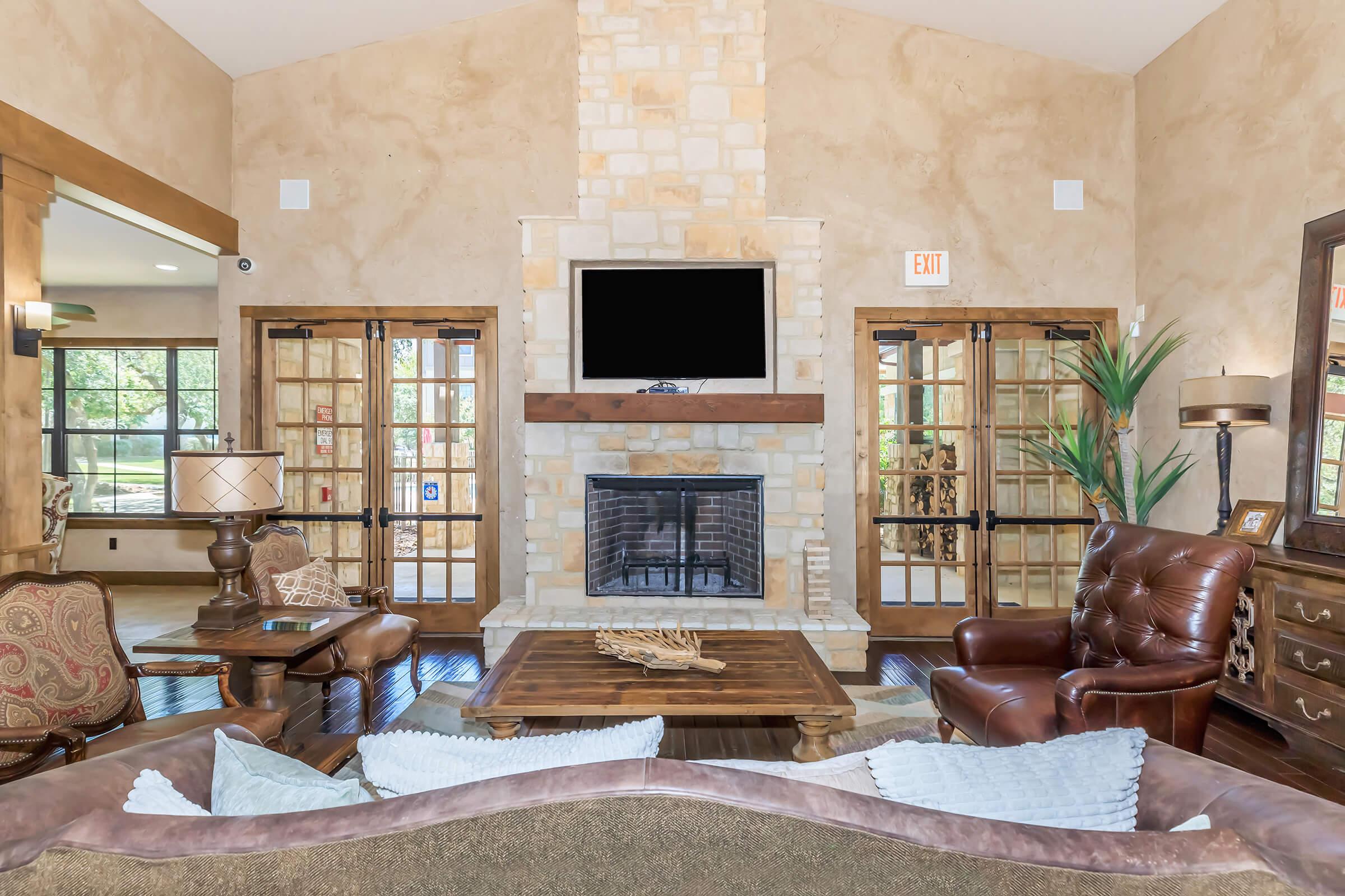 Cozy living room with a stone fireplace, mounted black TV, and large wooden coffee table. The space features two glass-paneled doors leading outside, a lamp beside a brown leather chair, and a decorative plant. Natural light filters in through the windows, enhancing the warm, inviting atmosphere.