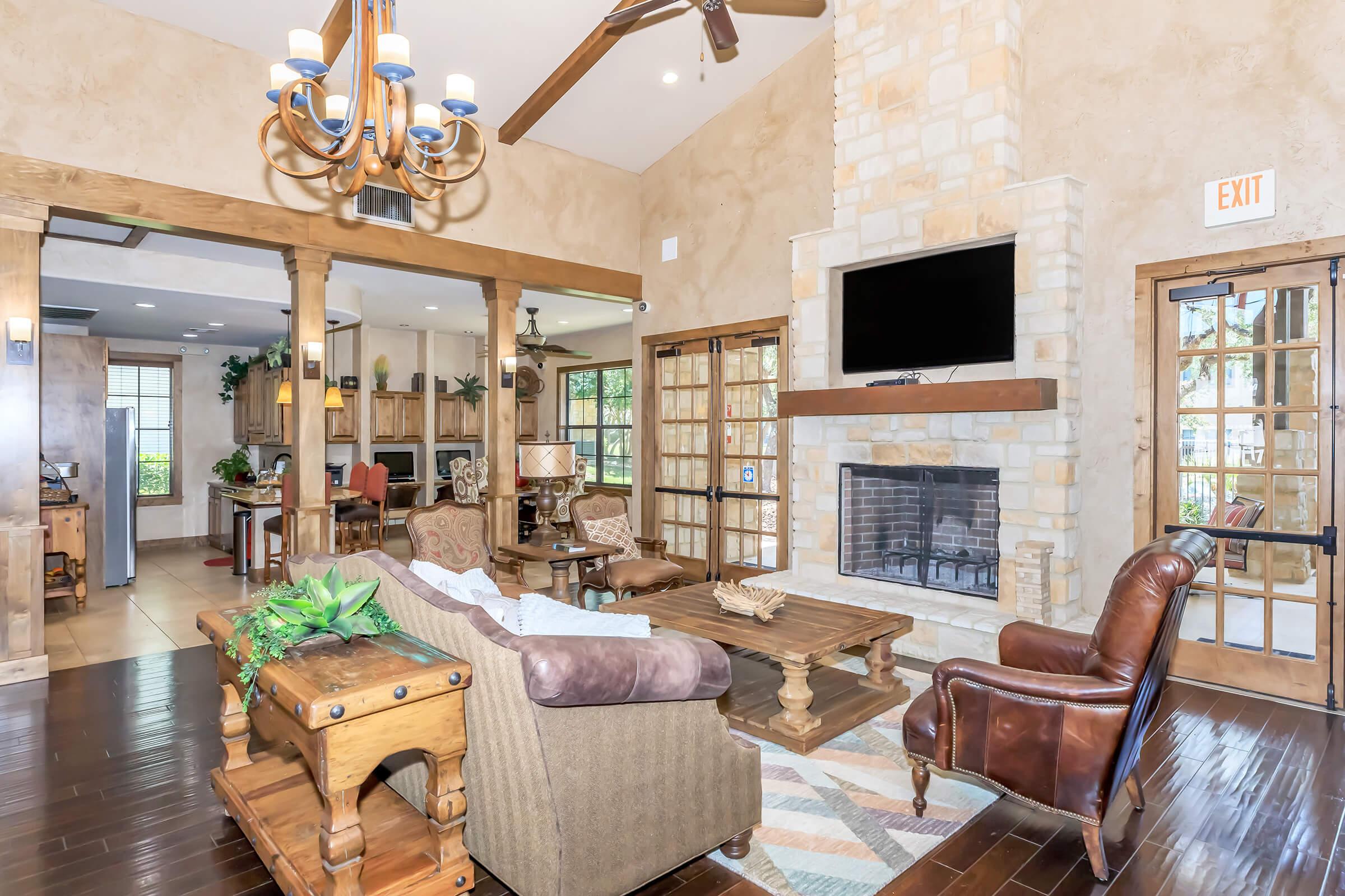 A cozy living room featuring a stone fireplace, dark wooden flooring, and a comfortable sofa. The space includes a wooden coffee table, decorative plants, and a chandelier hanging from the ceiling. Large windows provide natural light, and adjacent to the living area is a well-furnished kitchen visible through an open layout.