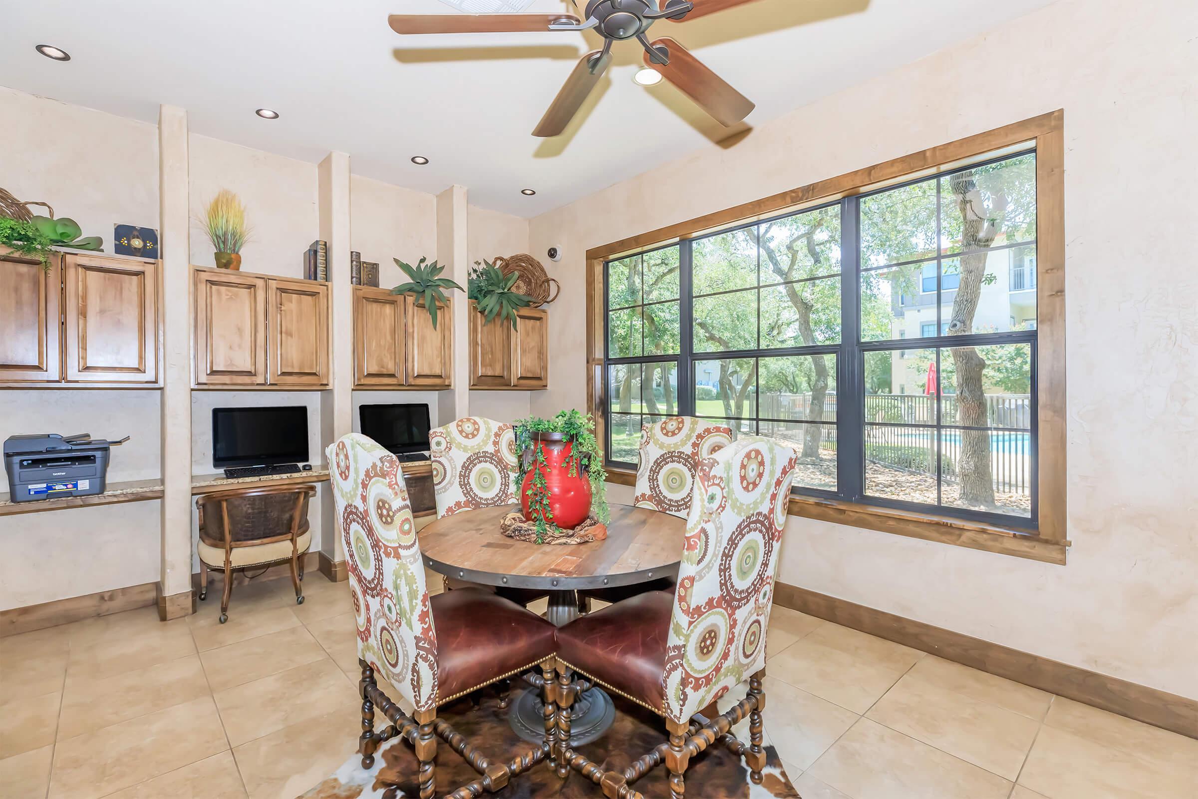 A cozy dining area with a round table surrounded by four upholstered chairs, featuring patterned fabric. There are built-in wooden cabinets in the background, and two computers. Large windows allow natural light to fill the space, highlighting greenery outside. The décor includes a vase and plants for a warm atmosphere.
