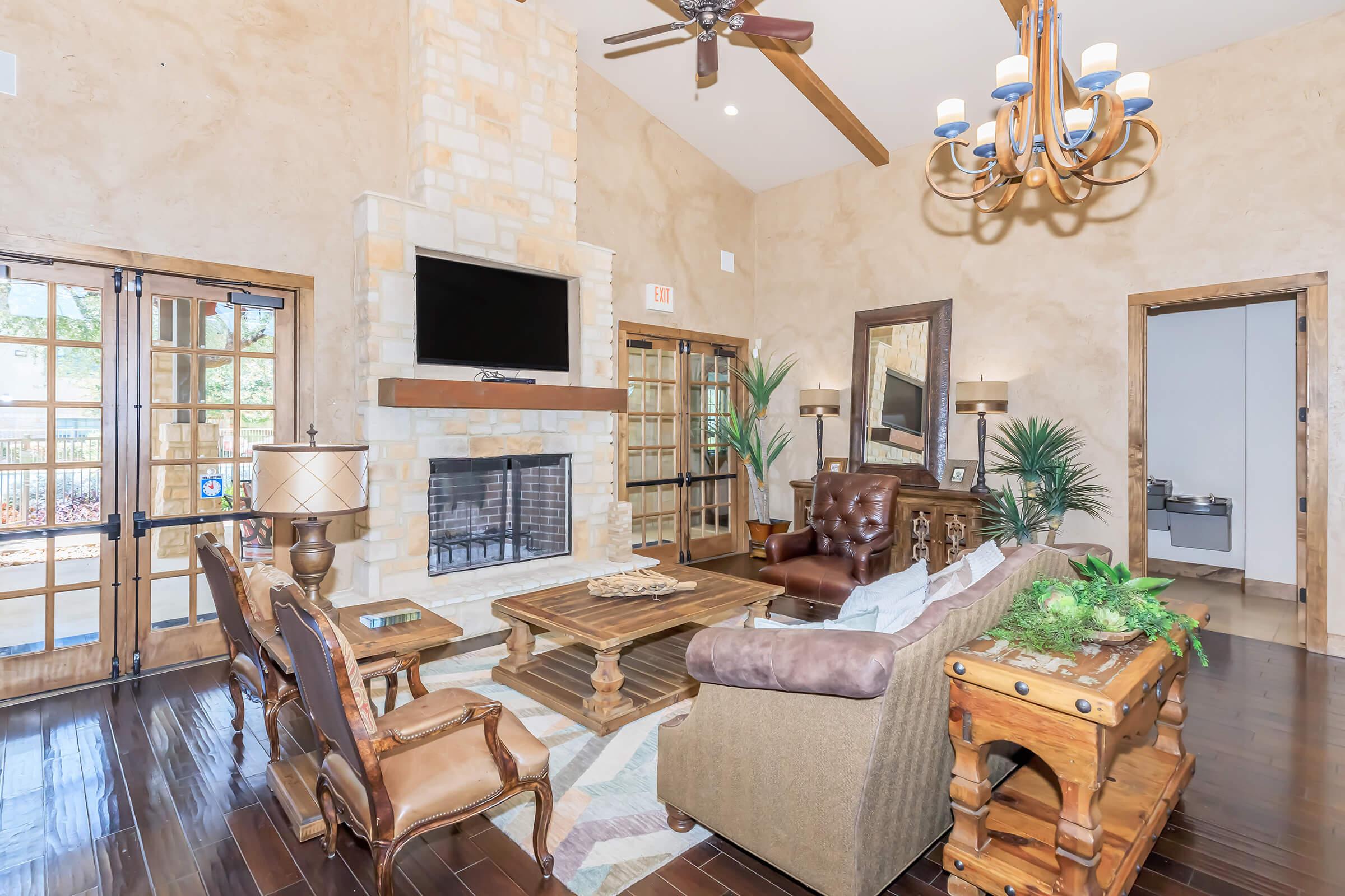 Cozy living room featuring a stone fireplace, a large TV mounted above it, and wooden floors. Comfortable seating includes a brown leather sofa and elegant armchairs. A wooden coffee table and decorative plants enhance the ambiance, with large windows and double doors allowing natural light to flow in.