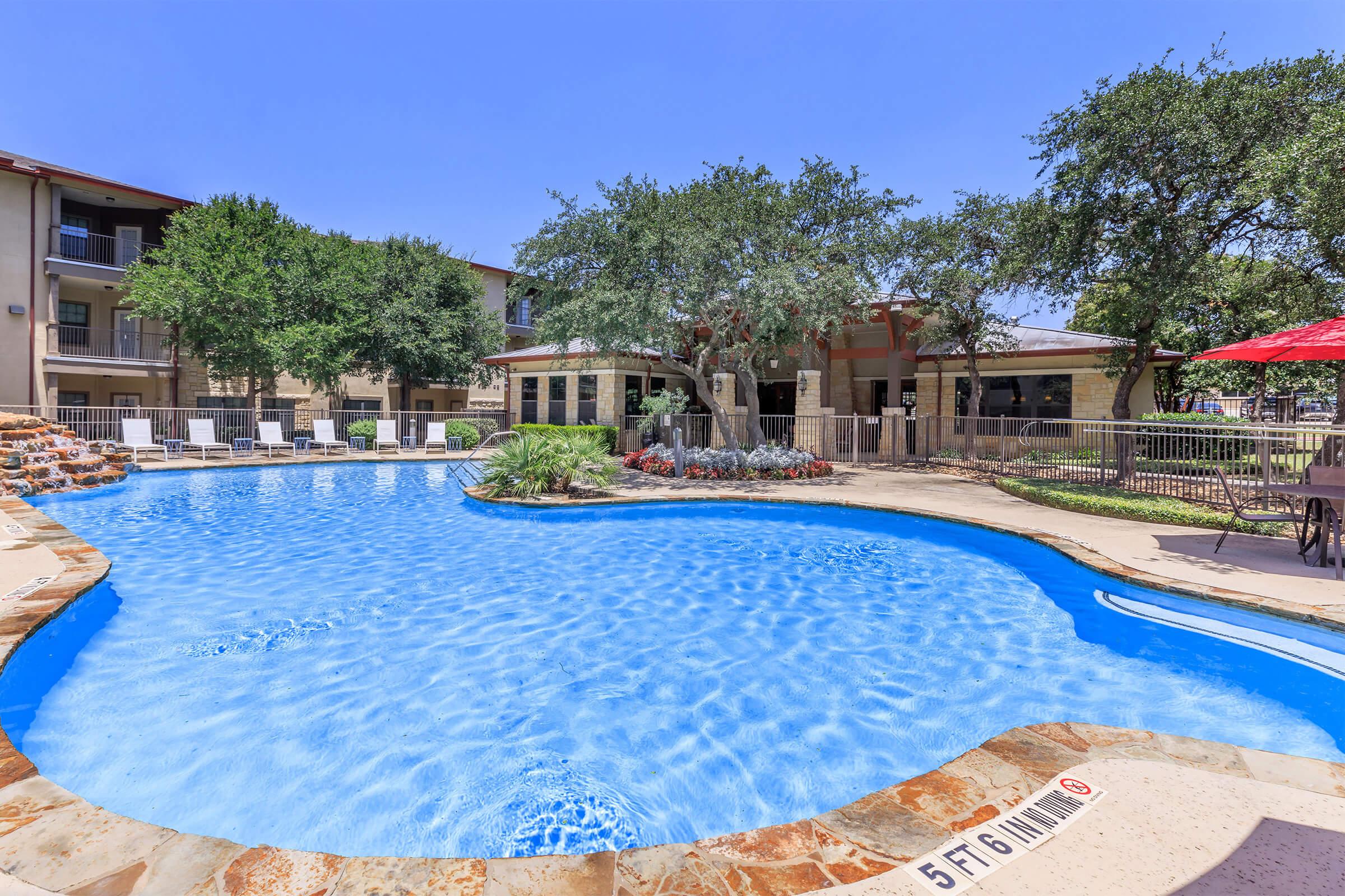 A beautiful swimming pool surrounded by lush greenery and lounge chairs, with a casual seating area and trees in the background. The pool has a vibrant blue color, and there’s a red umbrella nearby, creating a relaxing outdoor atmosphere.