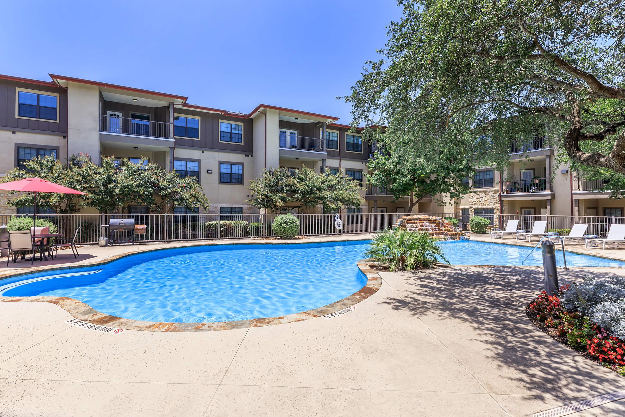 A clear blue swimming pool surrounded by lounge chairs and shaded areas, with lush greenery and blooming flowers nearby. Apartment buildings are visible in the background, creating a relaxing outdoor space for leisure and recreation.