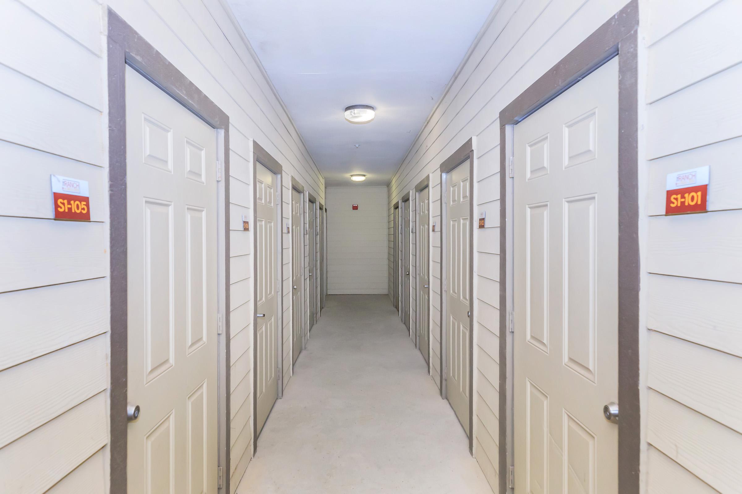 A well-lit corridor featuring several doors on either side, with room numbers displayed on plaques. The walls are light-colored wood paneling, and the floor is bare concrete. The space appears tidy and organized, suggesting a residential or storage facility.