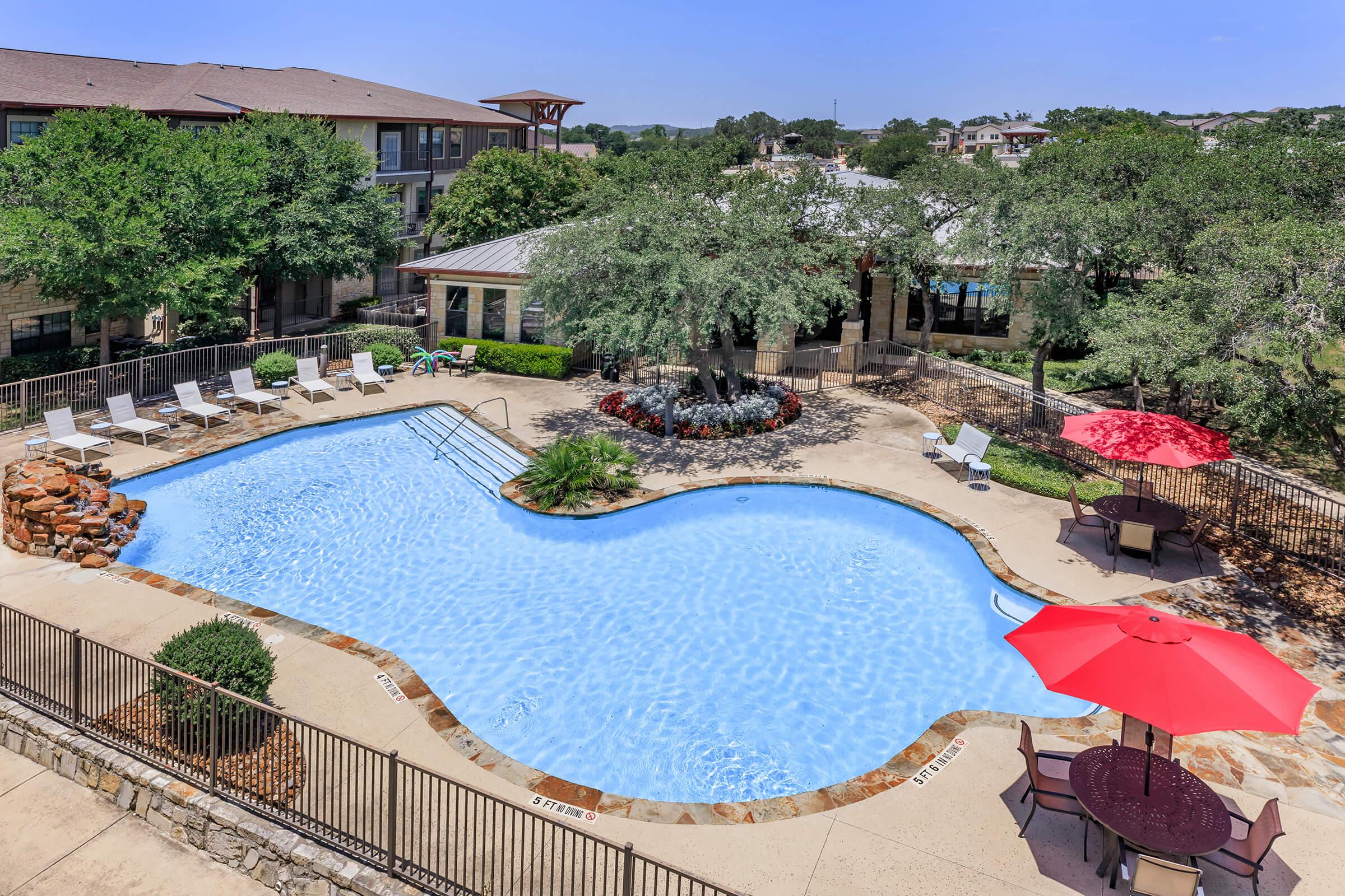 Aerial view of a serene swimming pool surrounded by lounge chairs and umbrellas. Lush greenery and decorative flower beds enhance the setting. Nearby, a shaded area with tables and chairs invites relaxation, while a building with large windows is visible in the background. Clear blue skies complete the picturesque scene.