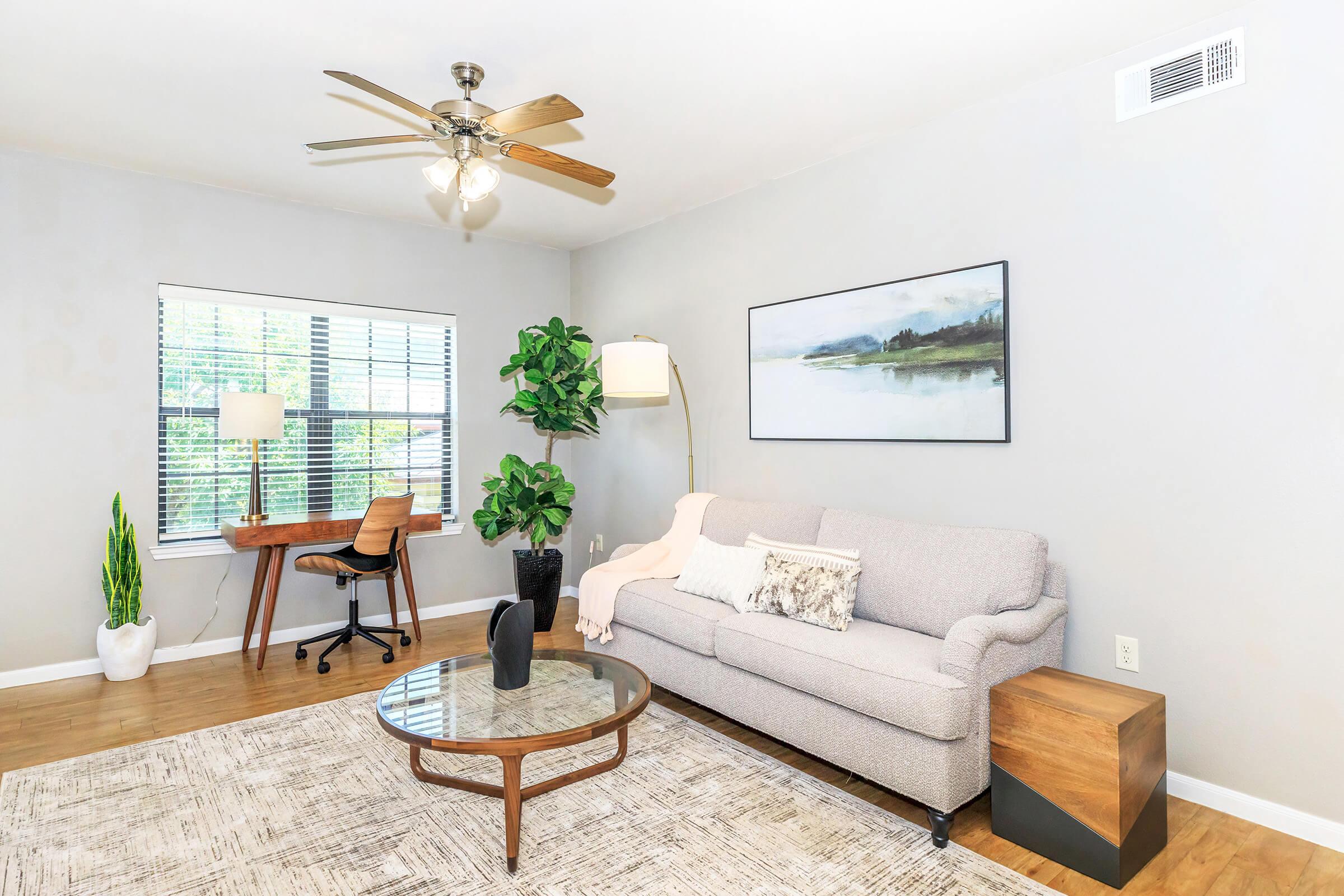 A cozy living room featuring a light gray sofa with decorative pillows, a wooden side table, and a round coffee table. There's a desk with a chair, a tall plant, and a wall-mounted lamp. A large window with blinds allows natural light, and a piece of artwork hangs above the sofa. The flooring is light wood.