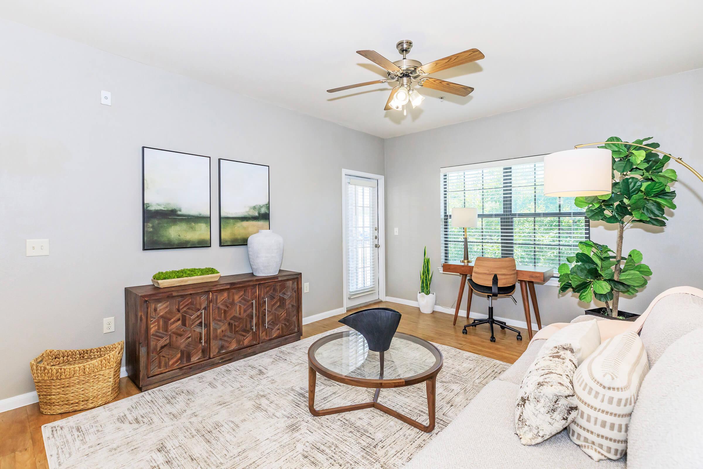 A modern living room featuring a light gray color scheme. It includes a comfortable sofa, a round glass coffee table, and a decorative wooden cabinet. A desk with a chair is positioned near a window, with greenery and a floor lamp enhancing the space. Natural light filters in, creating a warm atmosphere.