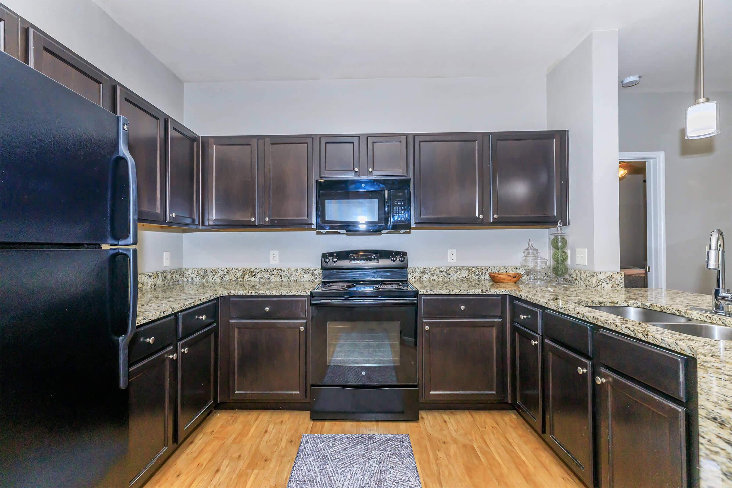 Modern kitchen featuring dark wooden cabinets, stainless steel appliances including a refrigerator and stove, a black microwave, and granite countertops. The flooring is a light-colored wood, and a runner rug adds a decorative touch. The kitchen is well-lit and organized, showcasing a spacious layout.
