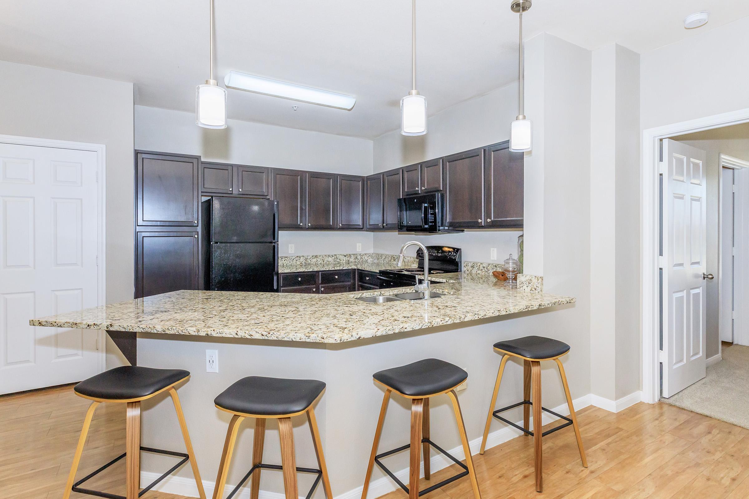 Modern kitchen with dark wooden cabinets, a granite countertop featuring a curved breakfast bar, and four minimalist stools. Appliances include a black refrigerator and a microwave. The space is well-lit with pendant lighting and has an open layout, connecting to a hallway with doors leading to other rooms.