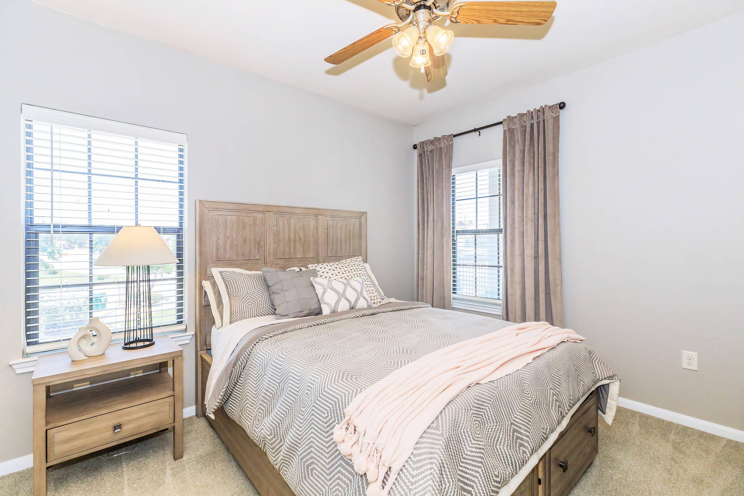 A cozy bedroom featuring a queen-sized bed with decorative pillows, a light gray blanket, bedside table with a lamp, and sheer curtains on two windows. The room has a ceiling fan and neutral paint, creating a warm and inviting atmosphere. The carpeted floor adds comfort.