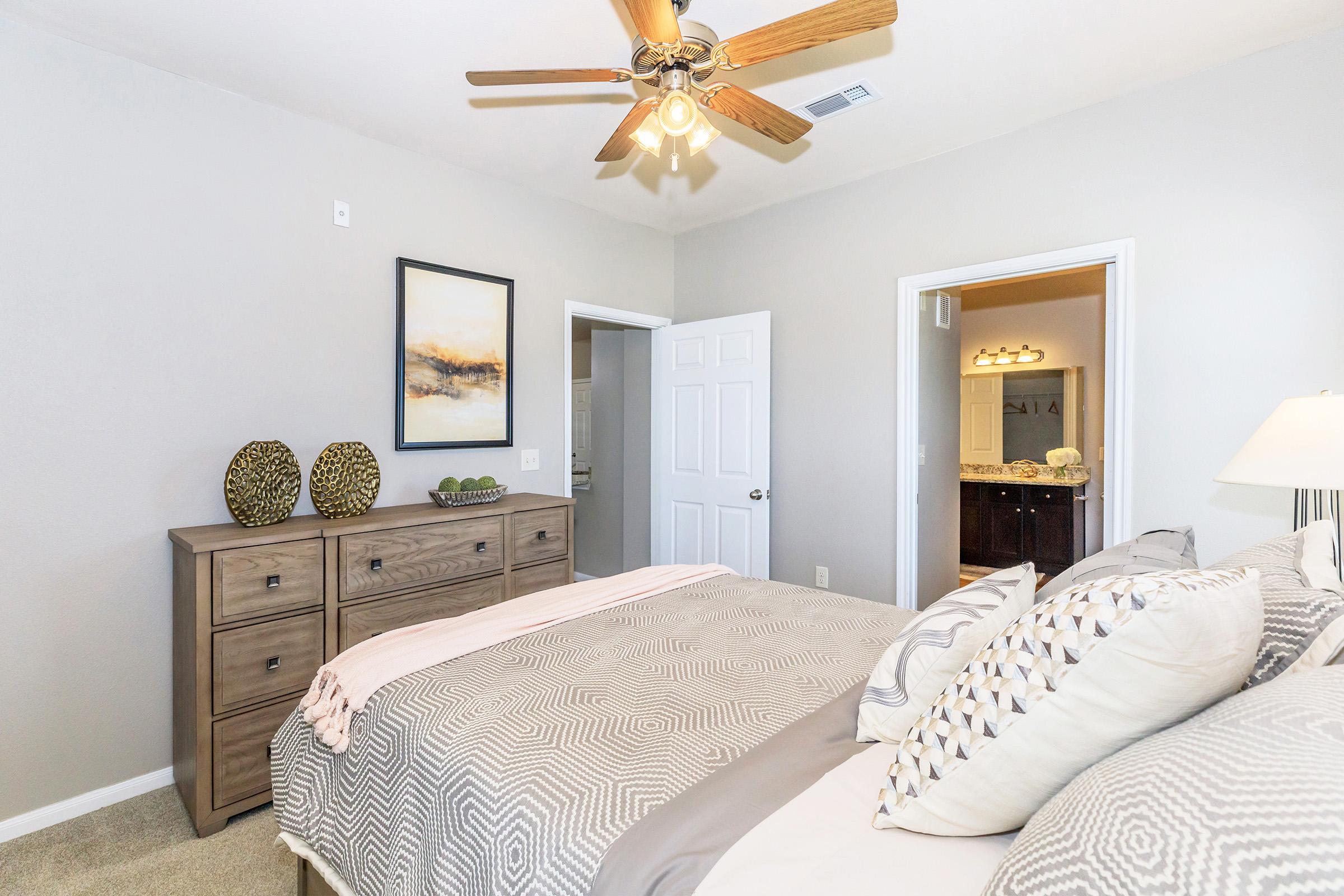 A cozy bedroom featuring a queen-sized bed with decorative pillows and a soft blanket. A wooden dresser is placed against the wall adorned with two decorative items. A ceiling fan is visible, and there’s a doorway leading to a bathroom, with neutral-colored walls enhancing the serene atmosphere.