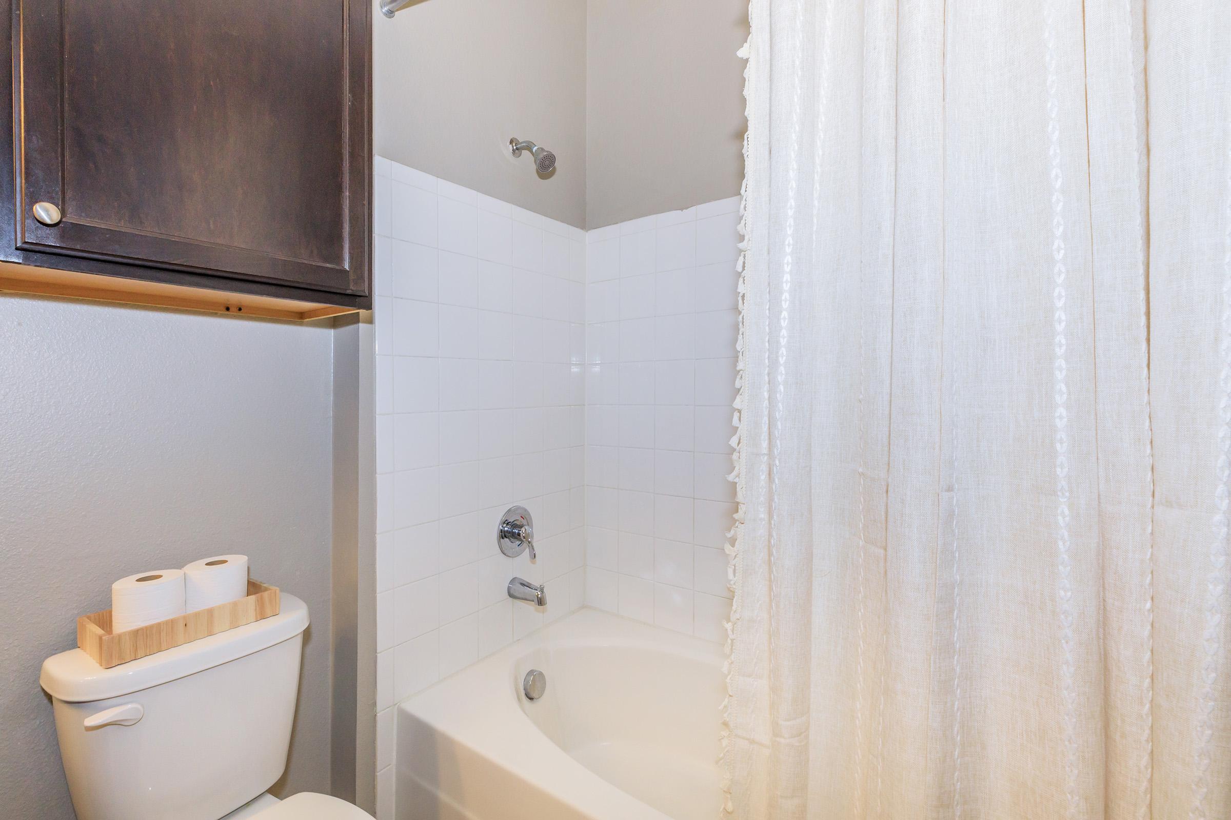 A clean bathroom featuring a bathtub with a shower head, a white shower curtain, a toilet with a small wooden shelf holding three rolls of toilet paper, and light gray walls. The space is well-lit and organized, highlighting a simple and modern design.