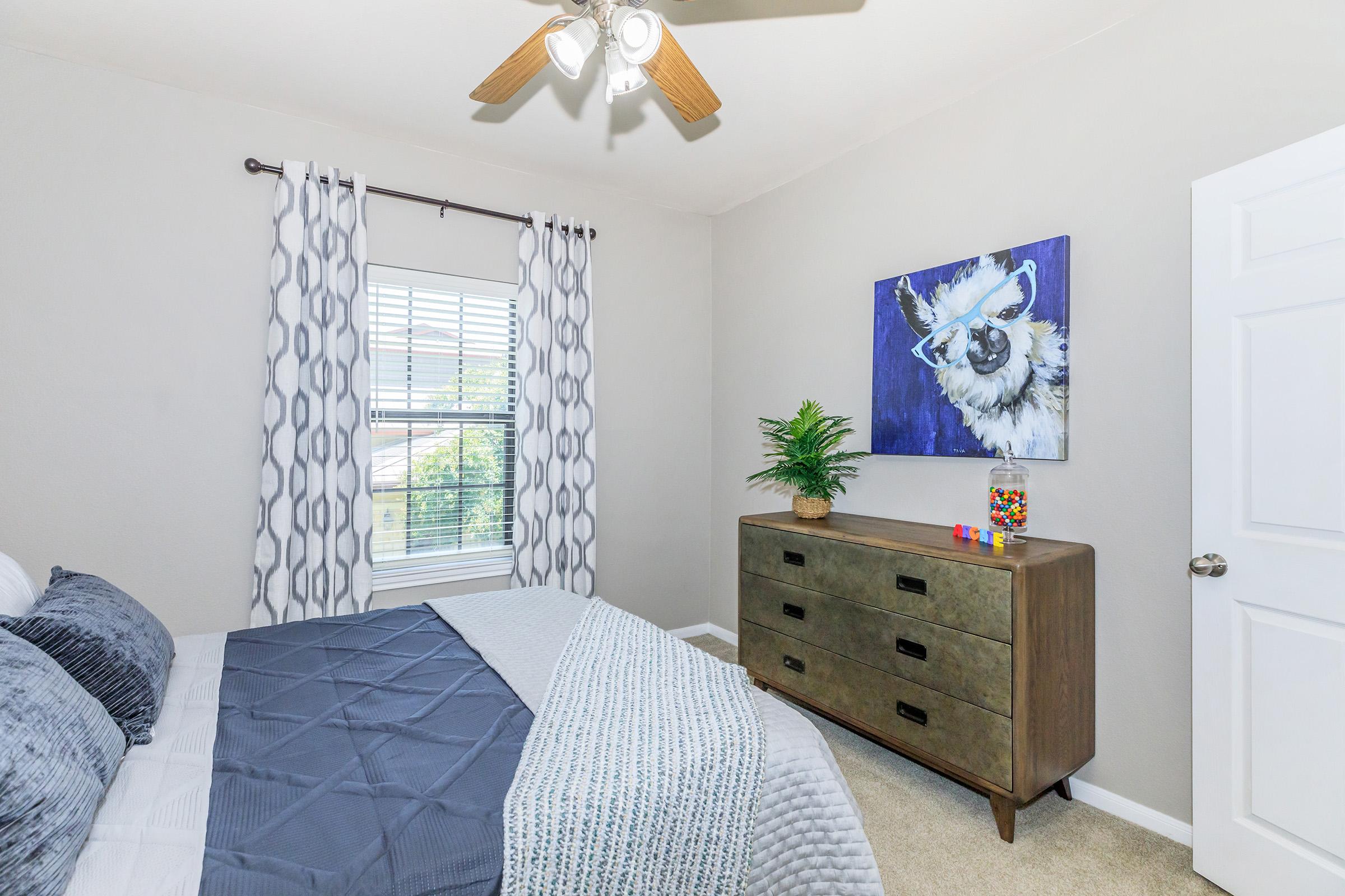 A cozy bedroom featuring a neatly made bed with gray and white linens, a wooden dresser with decorative items, and a colorful painting of a llama on the wall. Natural light enters through a window adorned with patterned curtains. The decor includes a small potted plant for added greenery.