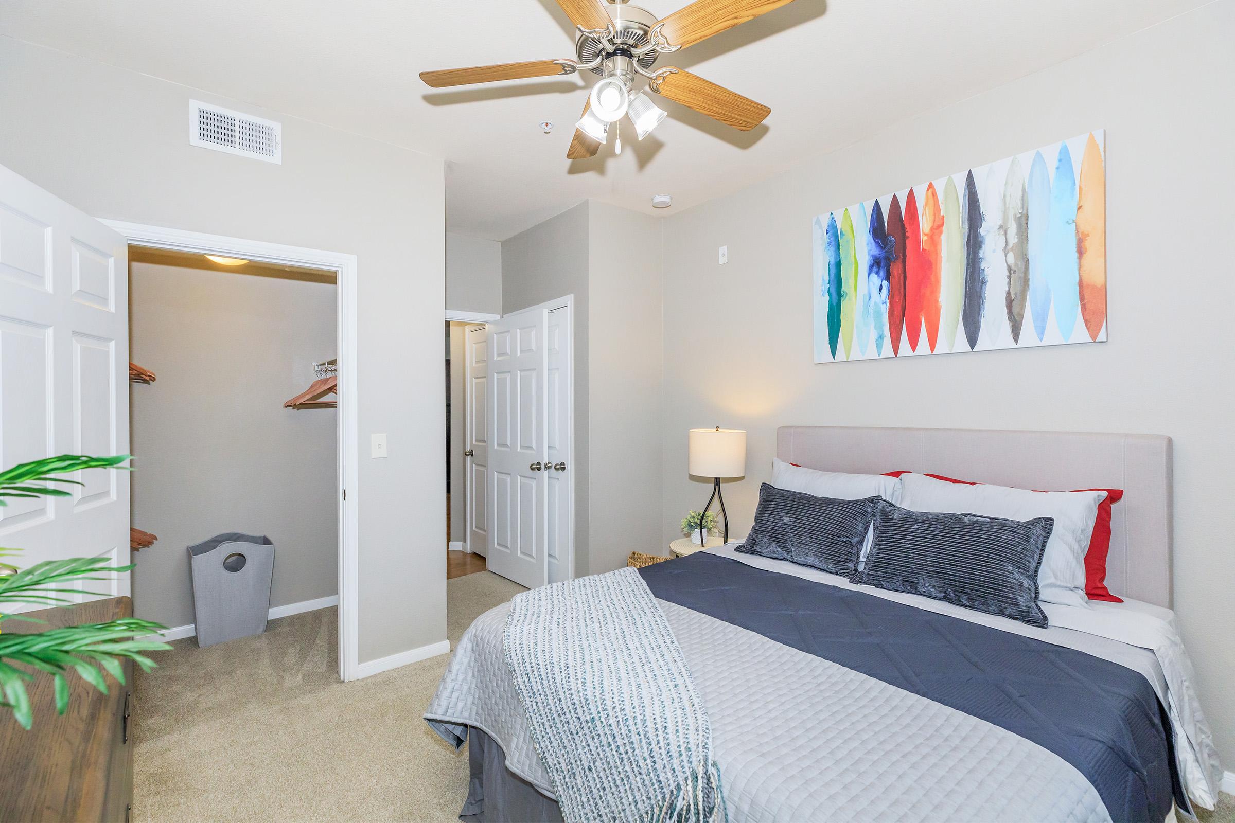 Cozy bedroom featuring a king-sized bed with gray and blue bedding, a bedside lamp, and a ceiling fan. An abstract painting in vibrant colors hangs above the headboard. To the left, a closet with wooden hangers is visible, and a laundry basket sits in the corner. The walls are painted in a soft gray tone.