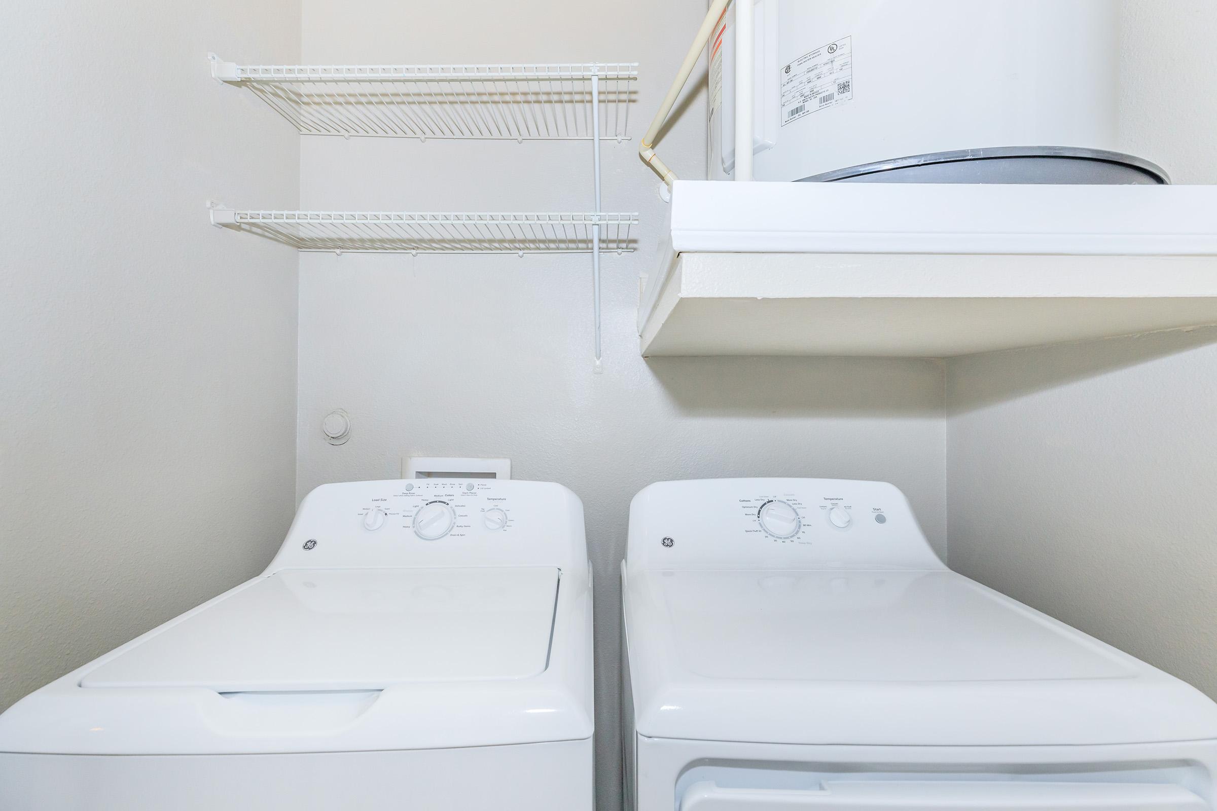 A laundry closet featuring a white washing machine and a dryer side by side. Above them is a white shelf for storage, and a water heater is partially visible on a shelf above the laundry appliances. The walls are painted a light color, creating a clean and organized appearance.