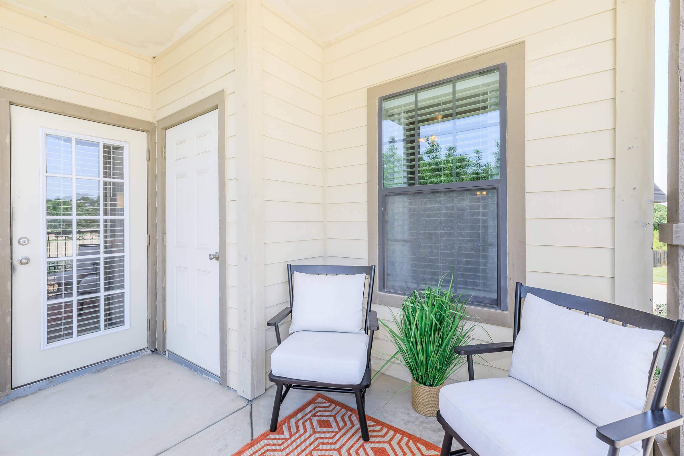 A cozy entryway featuring a small patio with two white chairs and a potted plant. The space has a decorative orange and white patterned rug, a door with glass panels, and a large window allowing natural light to brighten the area. The walls are painted light beige, creating a welcoming atmosphere.