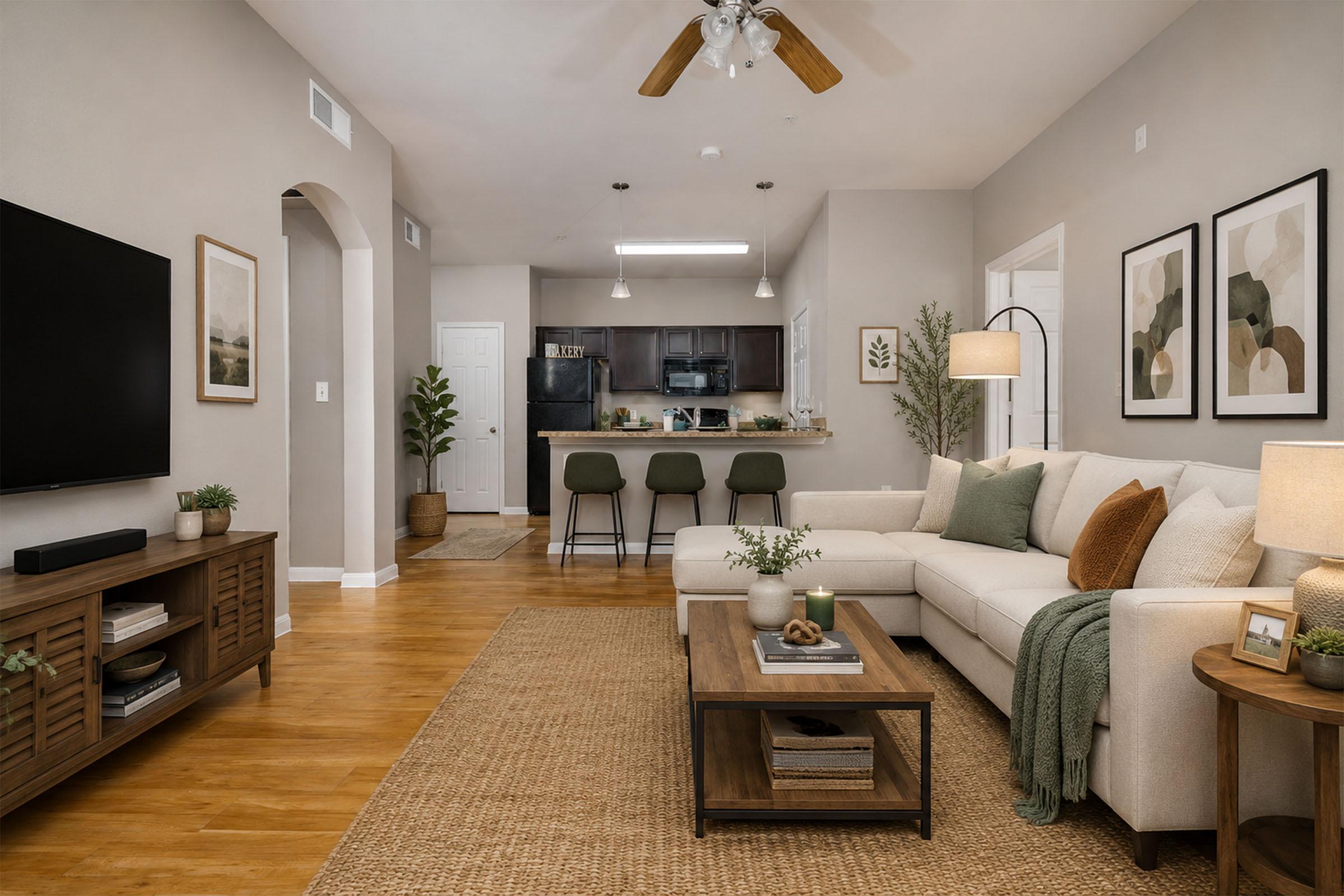 A modern living room featuring a light-colored sectional sofa with decorative pillows, a wooden coffee table, and a textured area rug. A TV is mounted on the wall opposite the couch. In the background, there's a kitchen with barstool seating and artwork on the walls, creating a cozy and inviting atmosphere.