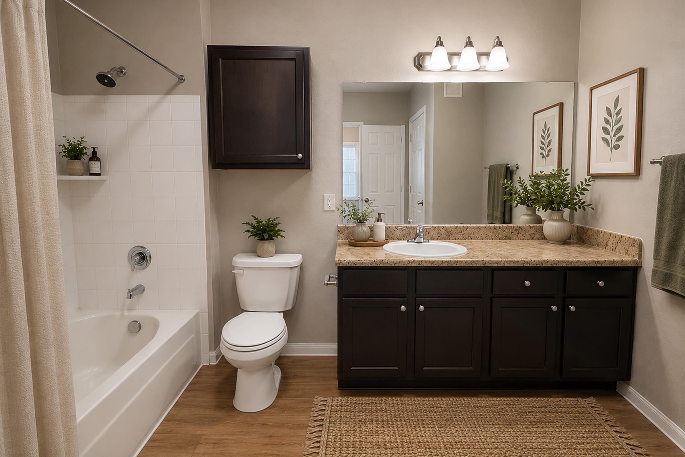 A clean, modern bathroom featuring a white tub with a shower curtain, a dark wooden vanity with a granite countertop, a round sink, and a mirror above it. There are potted plants on the counter, a toilet beside the vanity, and soft lighting from overhead fixtures. The floor is a light wood laminate, and the walls are painted a neutral color.