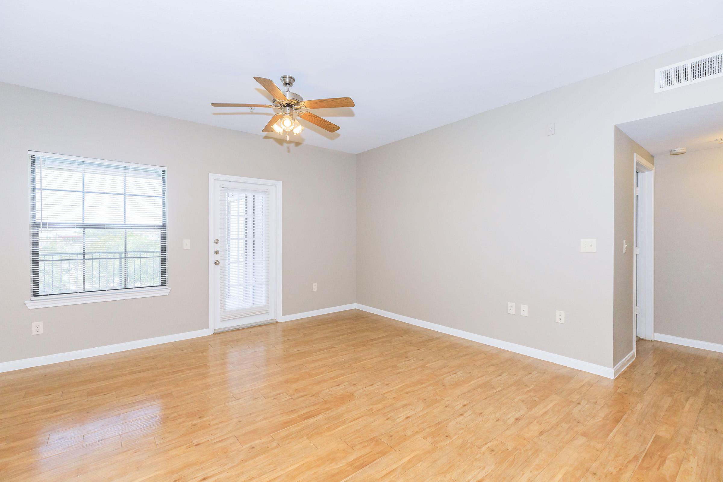 Spacious, empty living room featuring light hardwood floors, a ceiling fan, and large windows with blinds. A door leads to a balcony or outdoor area, while the walls are painted in a neutral color, creating a bright and airy atmosphere. Ideal for furnishing to personal taste.