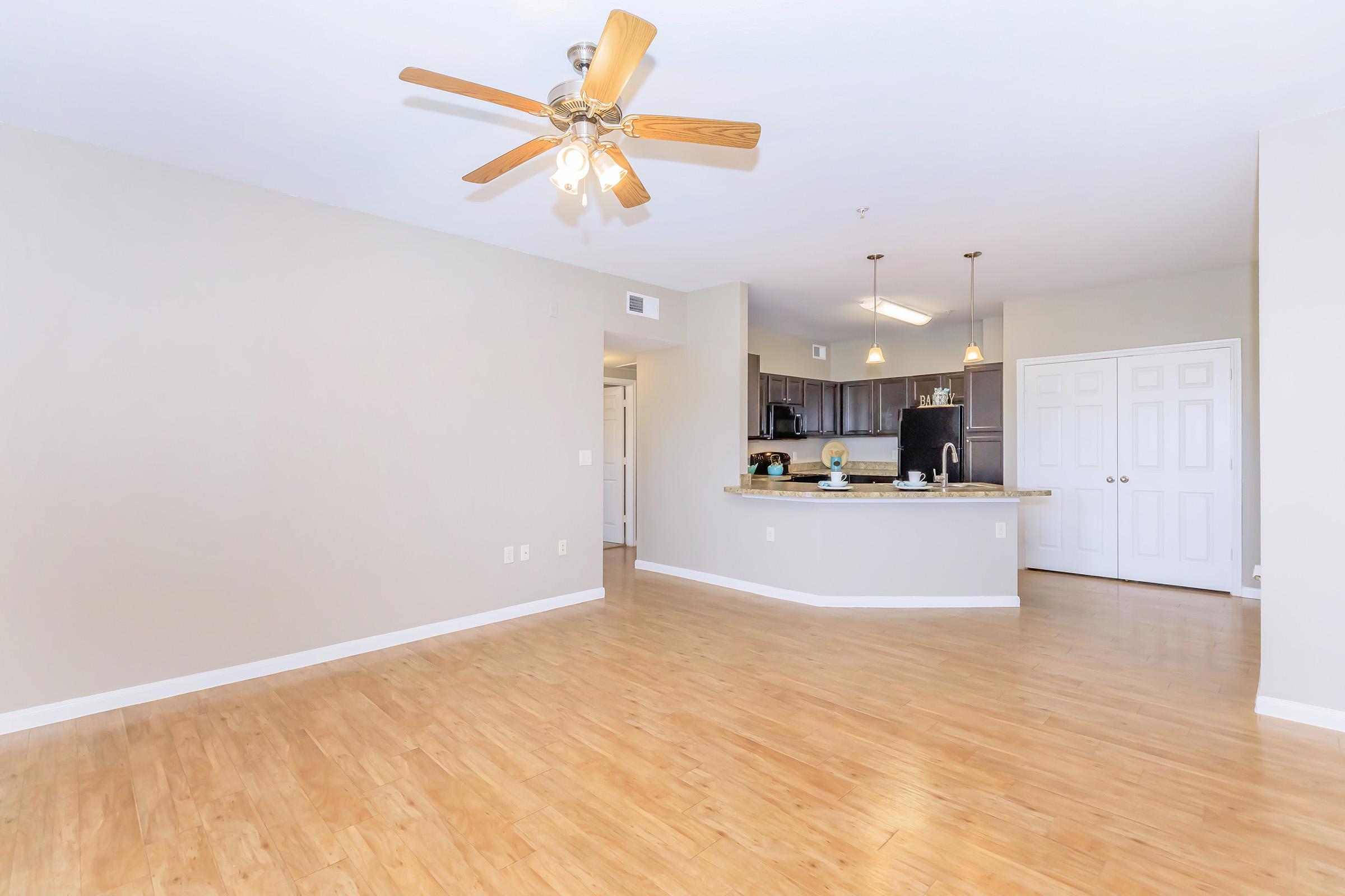 Spacious living area with light-colored wooden flooring, a ceiling fan, and neutral walls. The open layout connects to a modern kitchen featuring dark cabinetry and a countertop. Natural light brightens the space, enhancing the inviting atmosphere.