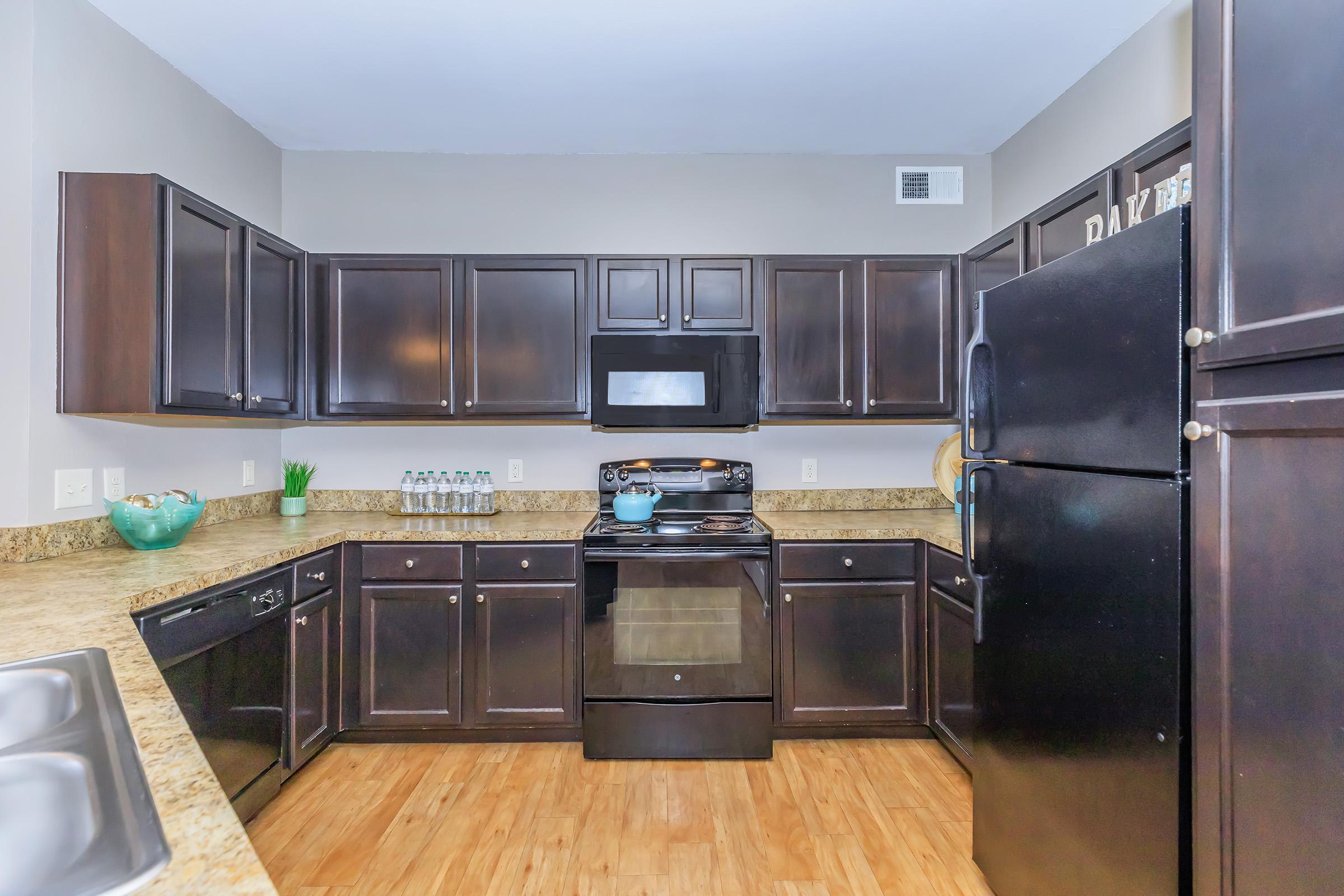 Modern kitchen featuring dark wooden cabinets, a black stove and microwave, granite countertops, and a black refrigerator. There's a window above the sink, and a vase with a plant on the counter. Bottled water is visible on the shelf. The flooring is a warm, light wood.