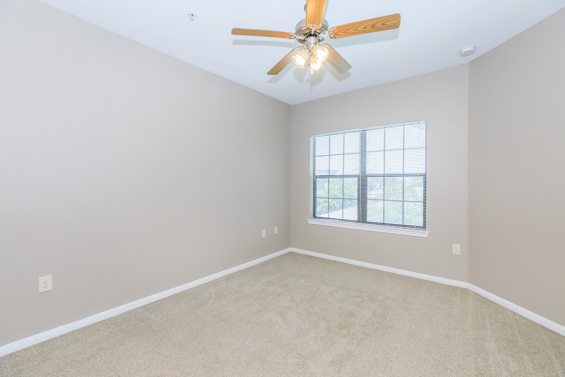 A vacant bedroom with light beige walls and carpet, featuring a ceiling fan with wooden blades and a light fixture. A large window allows natural light to enter, showcasing a view of the outdoors. The room has no furniture, creating a spacious and open feel.