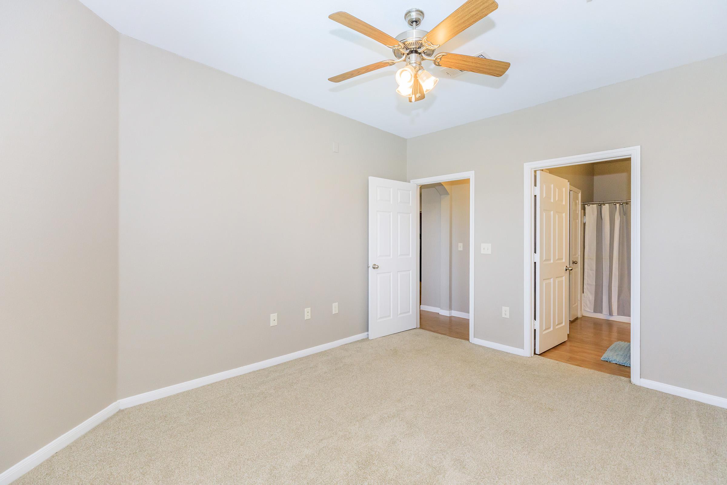 A spacious, empty bedroom featuring light beige walls and soft carpet. A ceiling fan with wooden blades hangs from the ceiling. Two doors lead to different areas, one possibly a closet and the other a bathroom, with a neutral color scheme creating a bright and airy atmosphere.