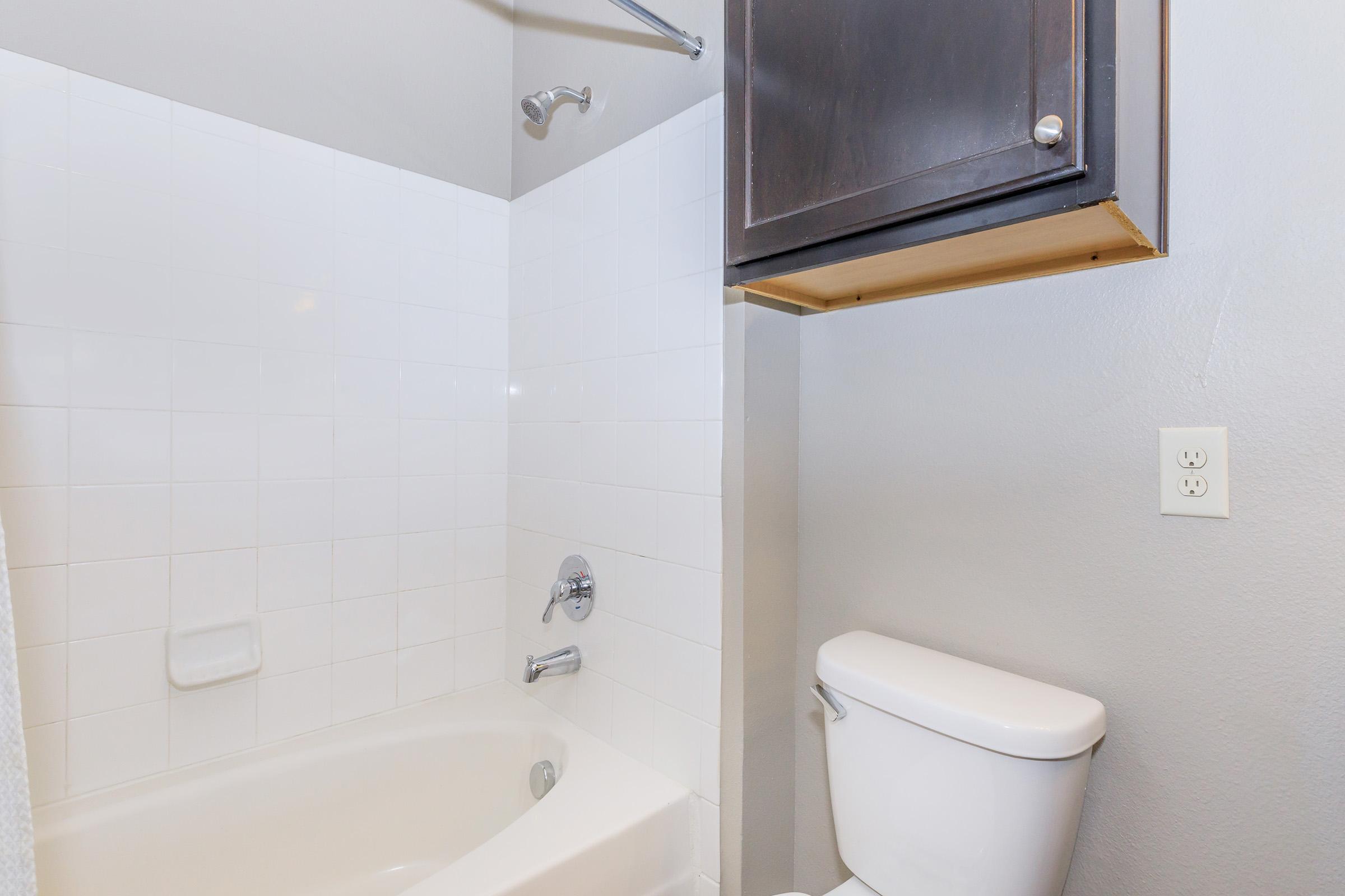 A clean bathroom featuring a white tiled bathtub with a showerhead, a modern toilet, and a small overhead cabinet above the tub. The walls are painted light gray, and there is an electrical outlet visible next to the toilet. The space appears bright and well-maintained.